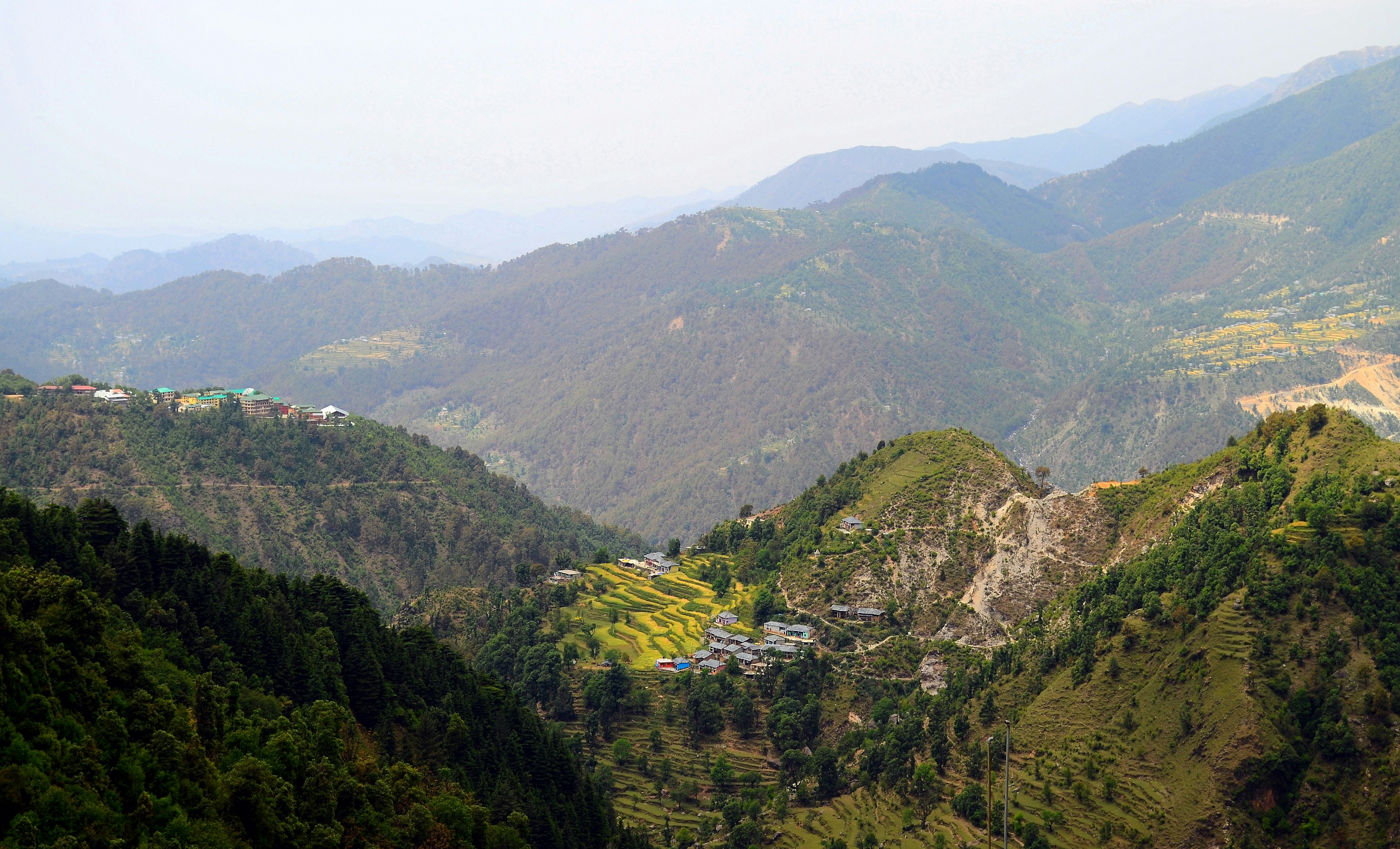 A sweeping view of the rolling hills of the Kangra Valley with a village on the side of a mountain visible in the foreground.