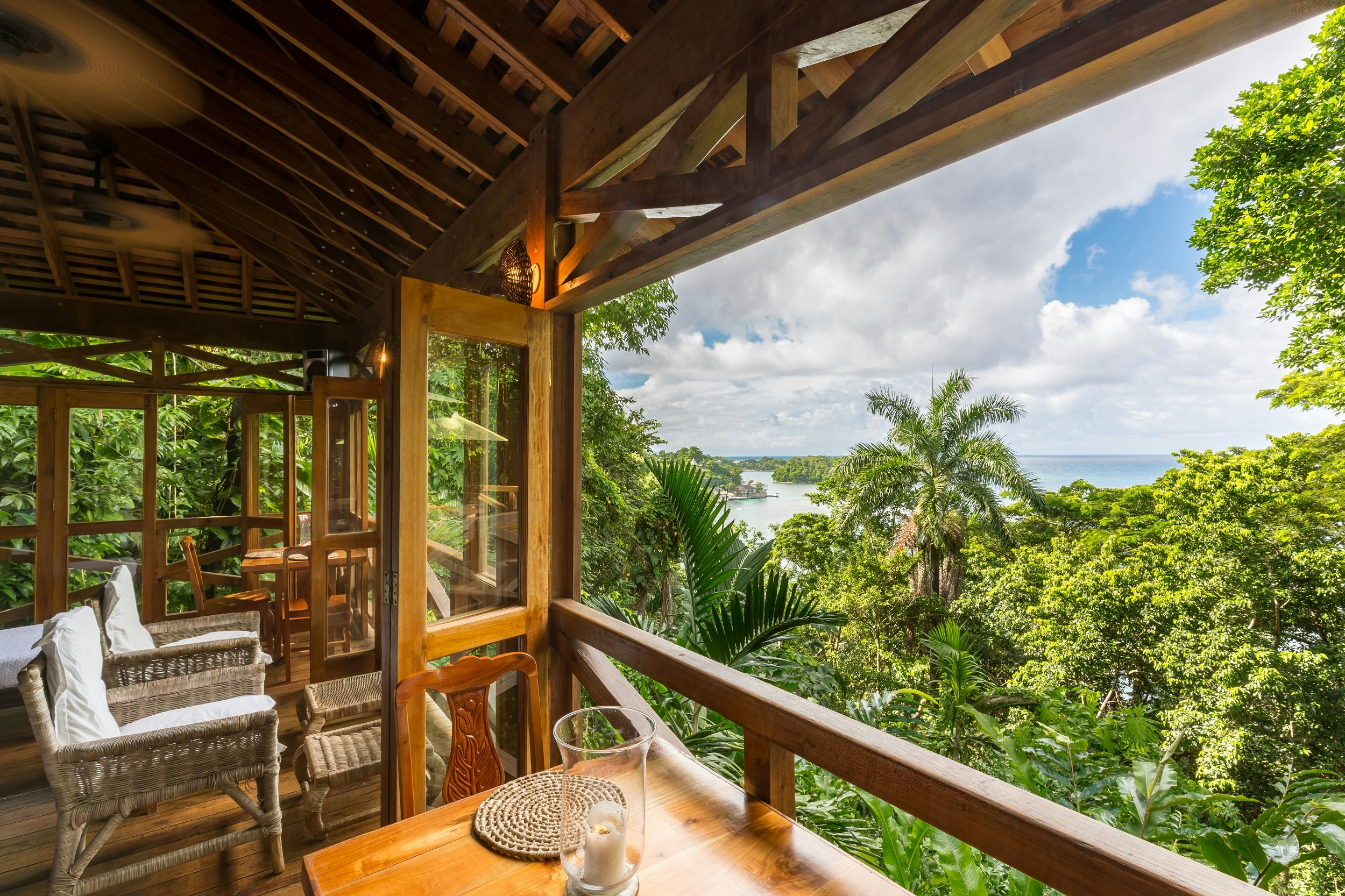 A view looking out through the open wall of Kanopi House's dining room; the wood-and-glass bifold doors open the entire wall of the traditional wood-framed structure and offer incredible views down over lush tropical forest to the coast below.