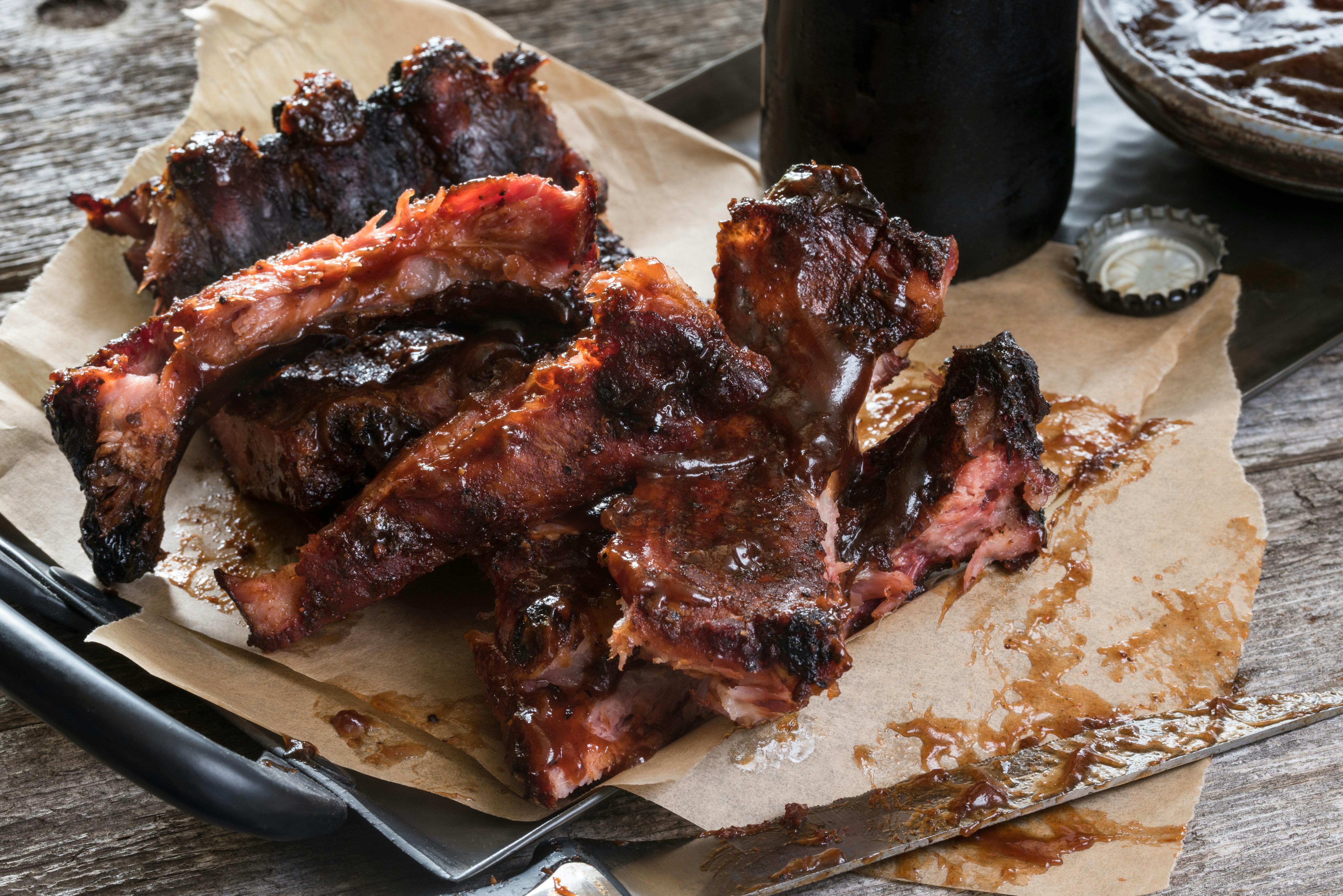 Closeup of a plate covered in light brown napkins and topped with a rack of smoked bbq. There's a knife covered in barbecue sauce to the right of the plate. At the top of the frame you can see the bottom section of a glass bottle. There is a bottle cap next to the bottle. 