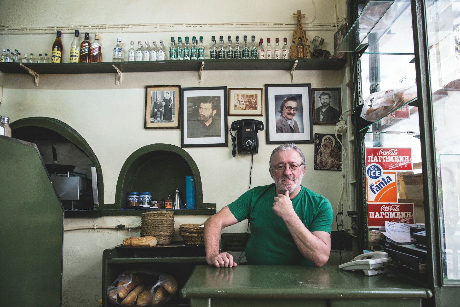 The proprietor of Kapetan Mixalis sits in a green shirt and glasses at the table of his Cretan-style taverna. Behind him are portraits of men with mid-century glasses and dramatic mustaches, as well as a vintage phone. Above on a high shelf are bottles of scotch and ouzo 