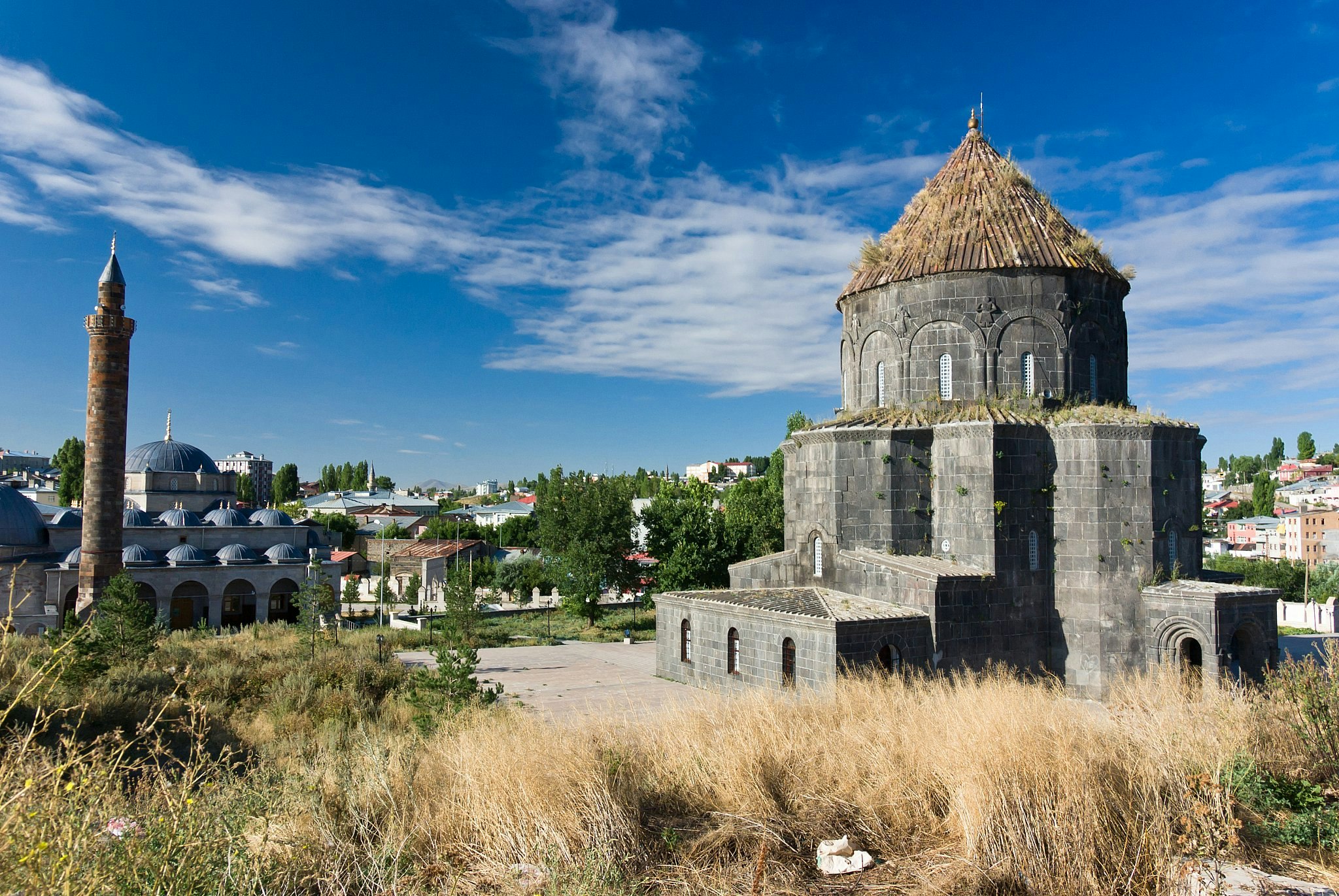 An imposing basalt church with a conical dome and porches below; next to it is a mosque with blue domes and a minaret.