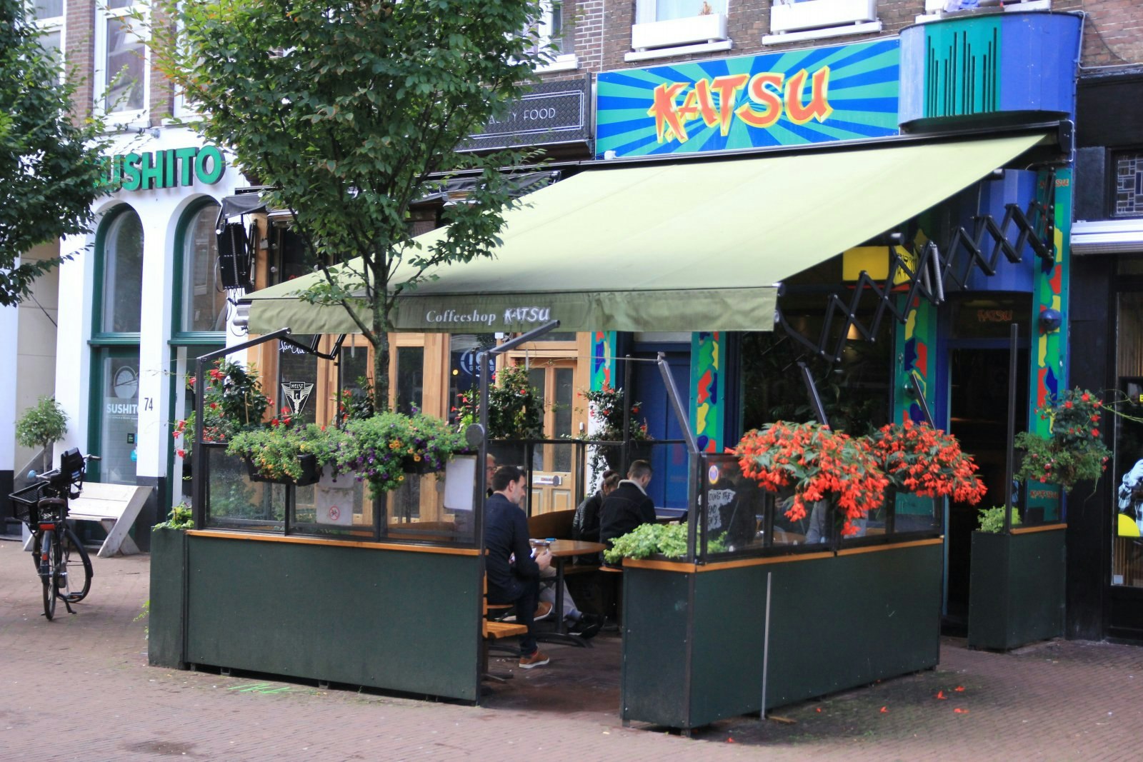 The exterior of Katsu coffeeshop in Amsterdam. The exterior is painted in vibrant colours and has a covered outdoor seating area surrounded by barriers and flowers. There are a few people in the seating area. 