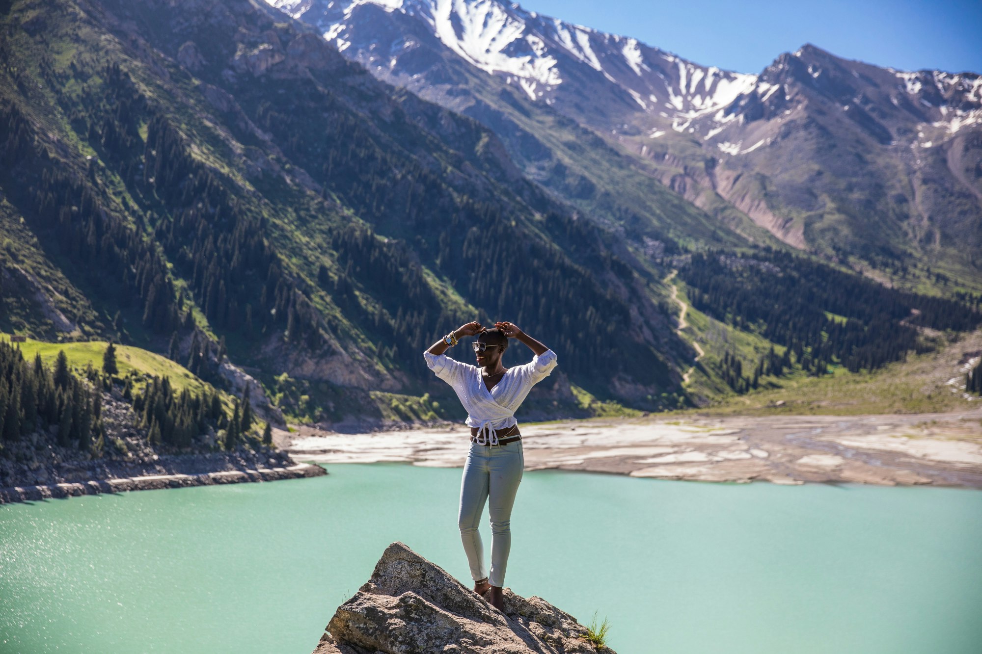 Jessica Nabongo smiles standing on a rock in front of a lake and green mountains in Kazakhstan