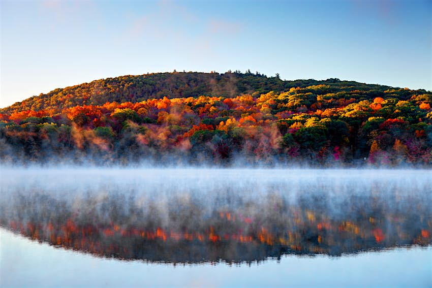 mist clings to the surface of a lake in Connecticut as red, orange, yellow and green leaves are reflected in its surface from a hill behind. New England fall foliage road trip