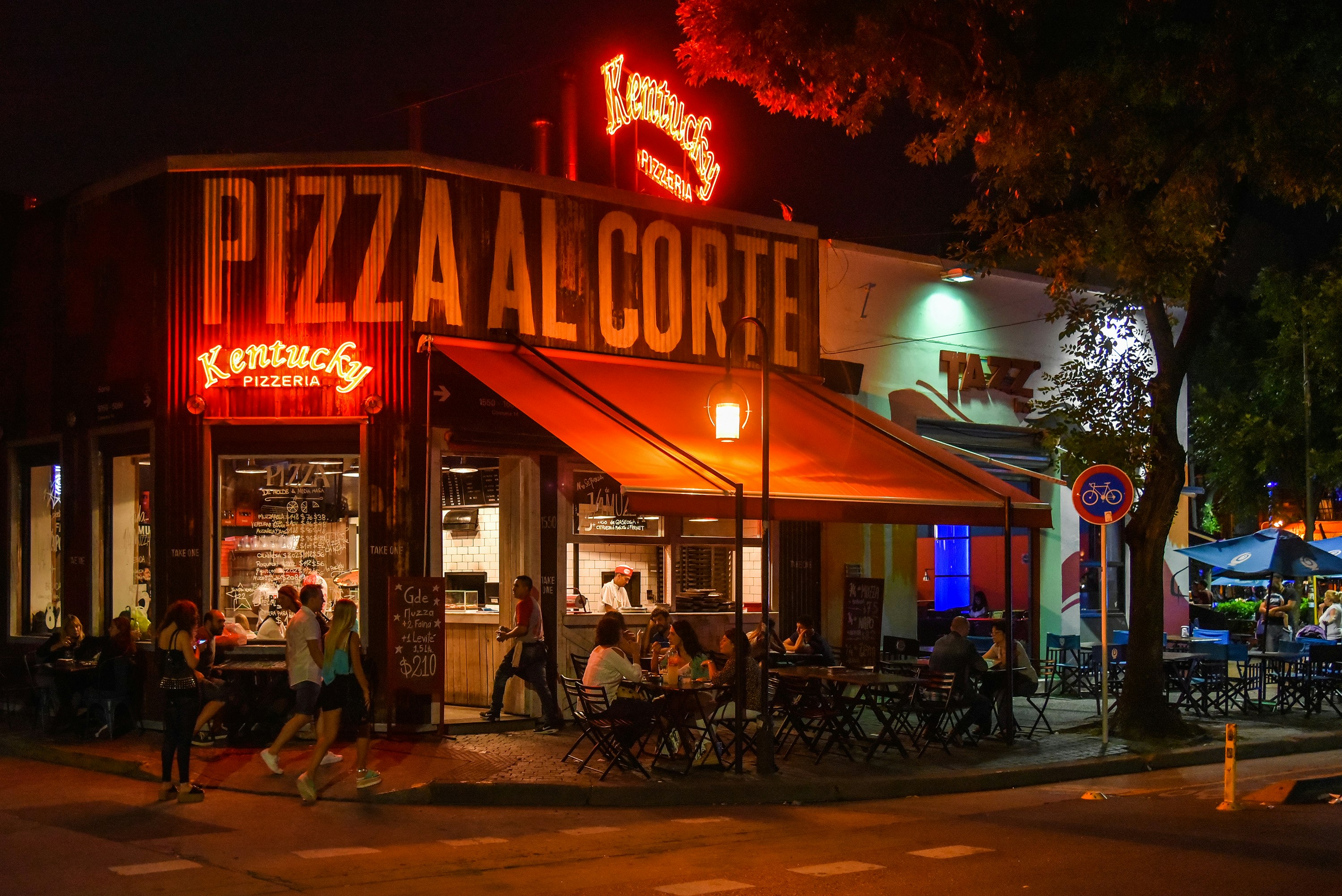 The red neon lights of Kentucky Pizzeria illuminate the street corner as people dine outside while others walk by  