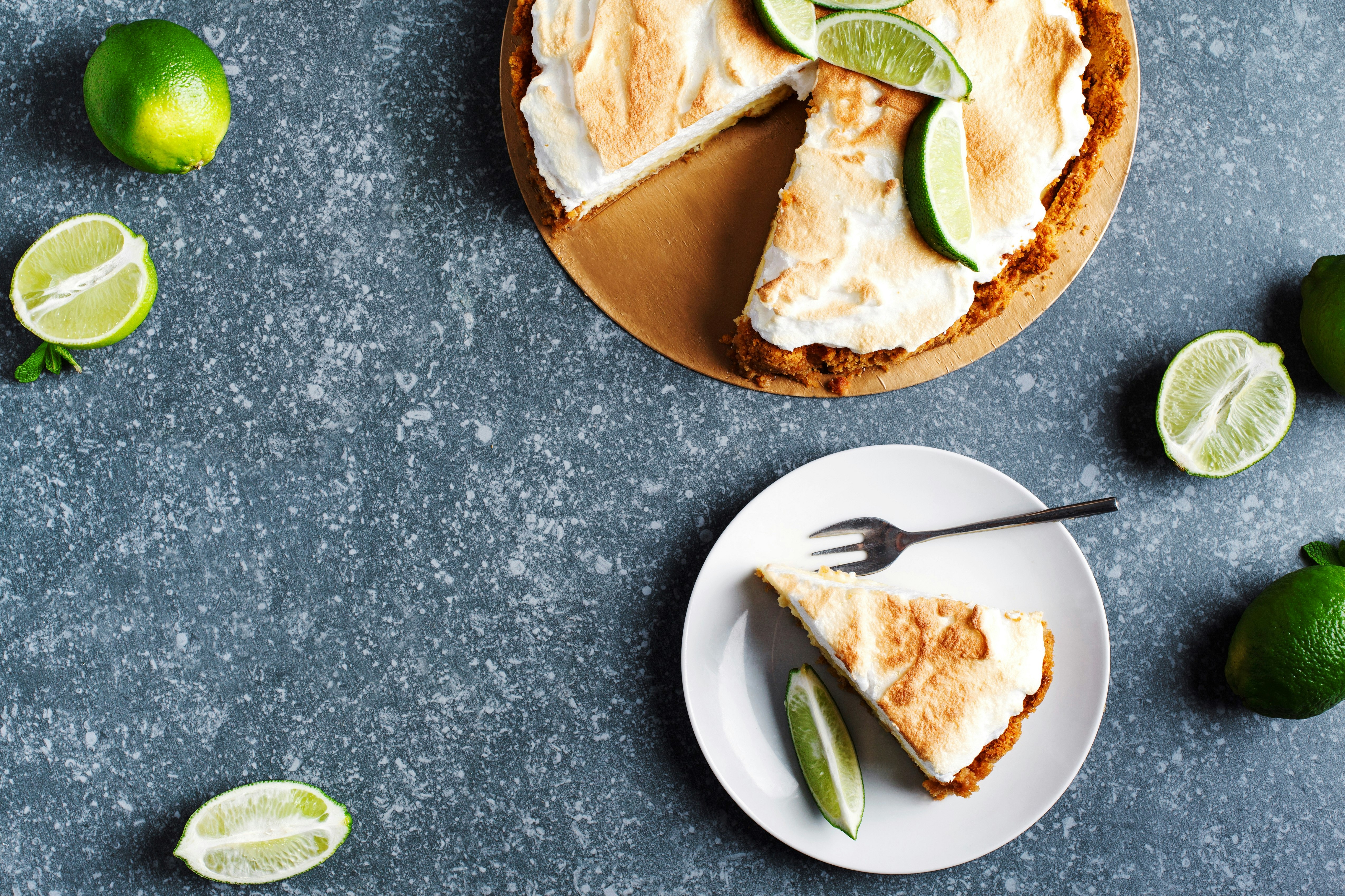 A wedge of a lime serves as garnish next to a slice of key lime pie. There is a fork at the top of the small plate. A few limes (some sliced into sections) are positioned around the plate and the full pie. 
