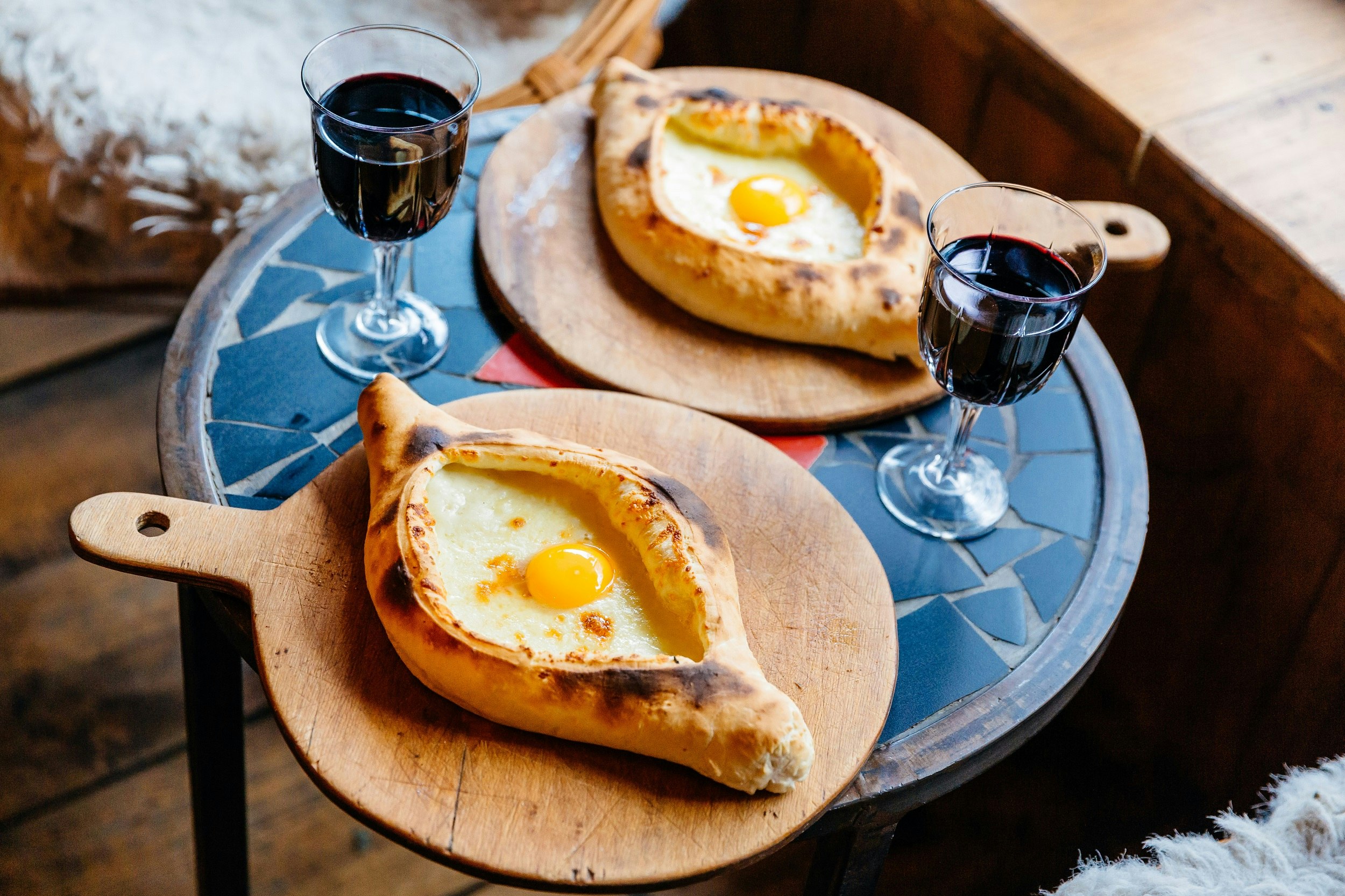 Two dishes of boat-shaped bread filled with cheese and egg, served on wooden boards with glasses of red wine.