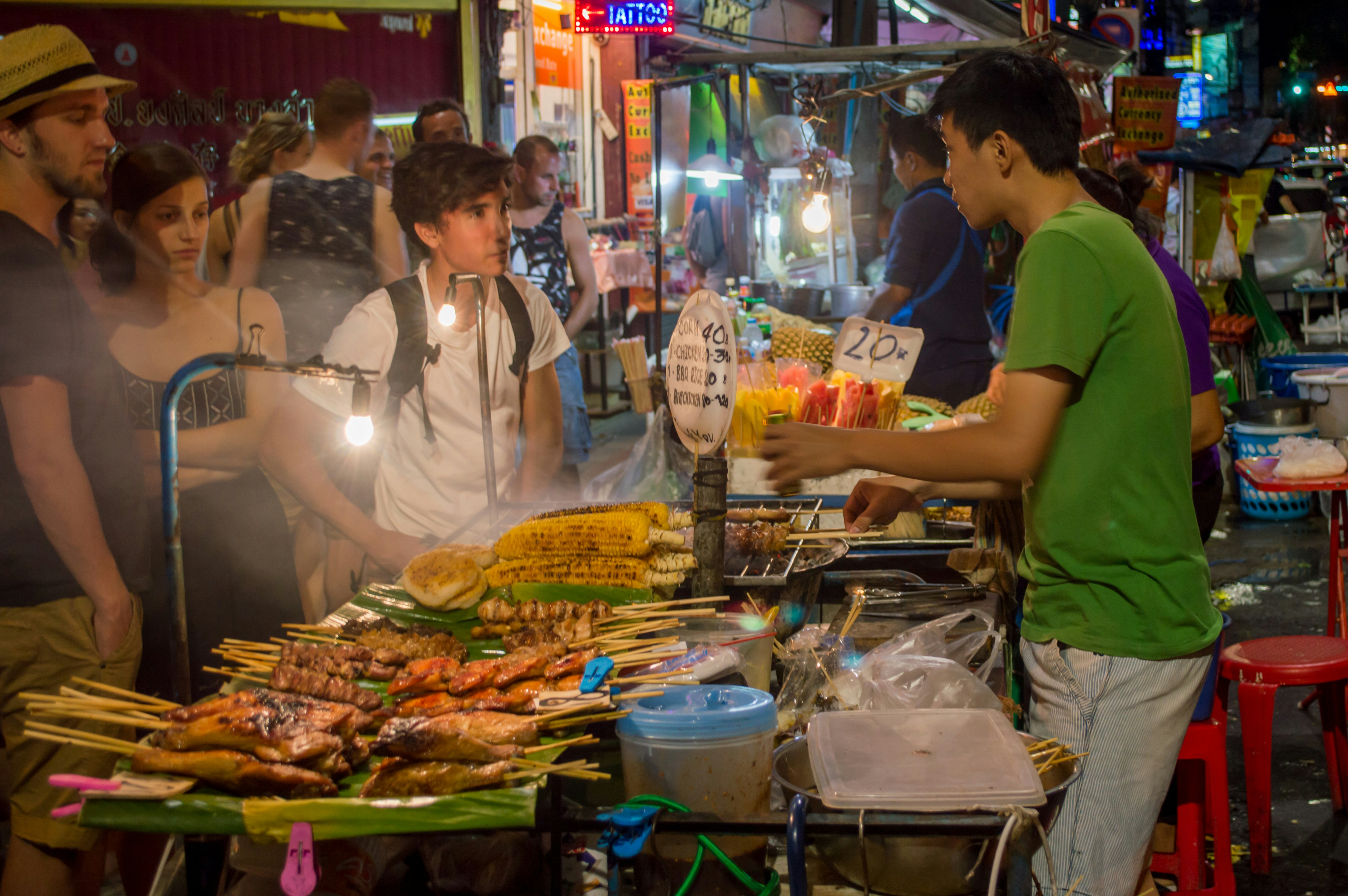 A street food stall on Bangkok's Khao San Road, where a vendor cooks kebabs and vegetables on a grill over an open flame, while a handful of tourists browse the offerings.