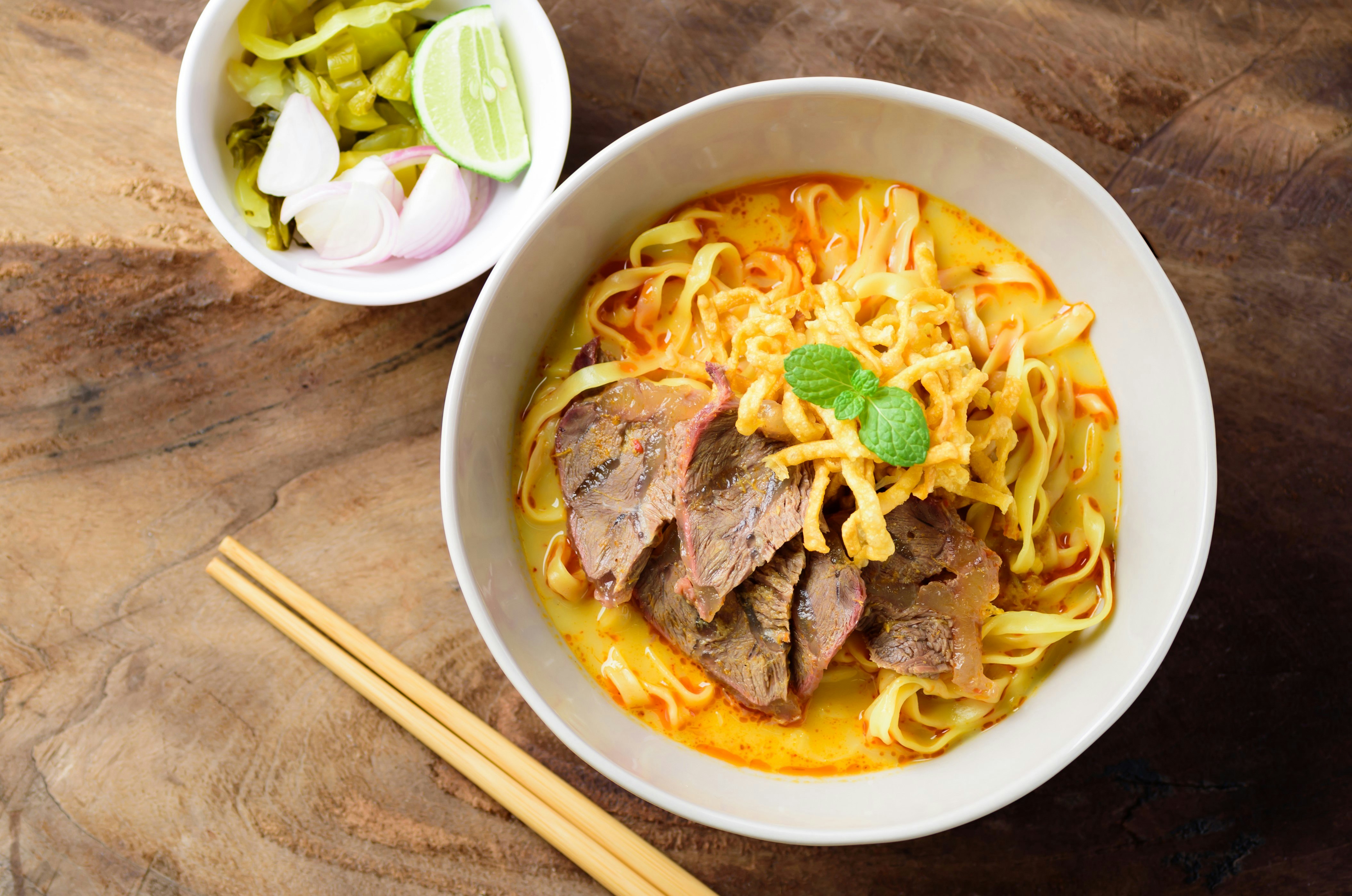 A top-down view of a bowl of khao soi sitting on a tabletop. Khao soi is a noodle soup with meat and vegetables with a creamy, yellowish broth.
