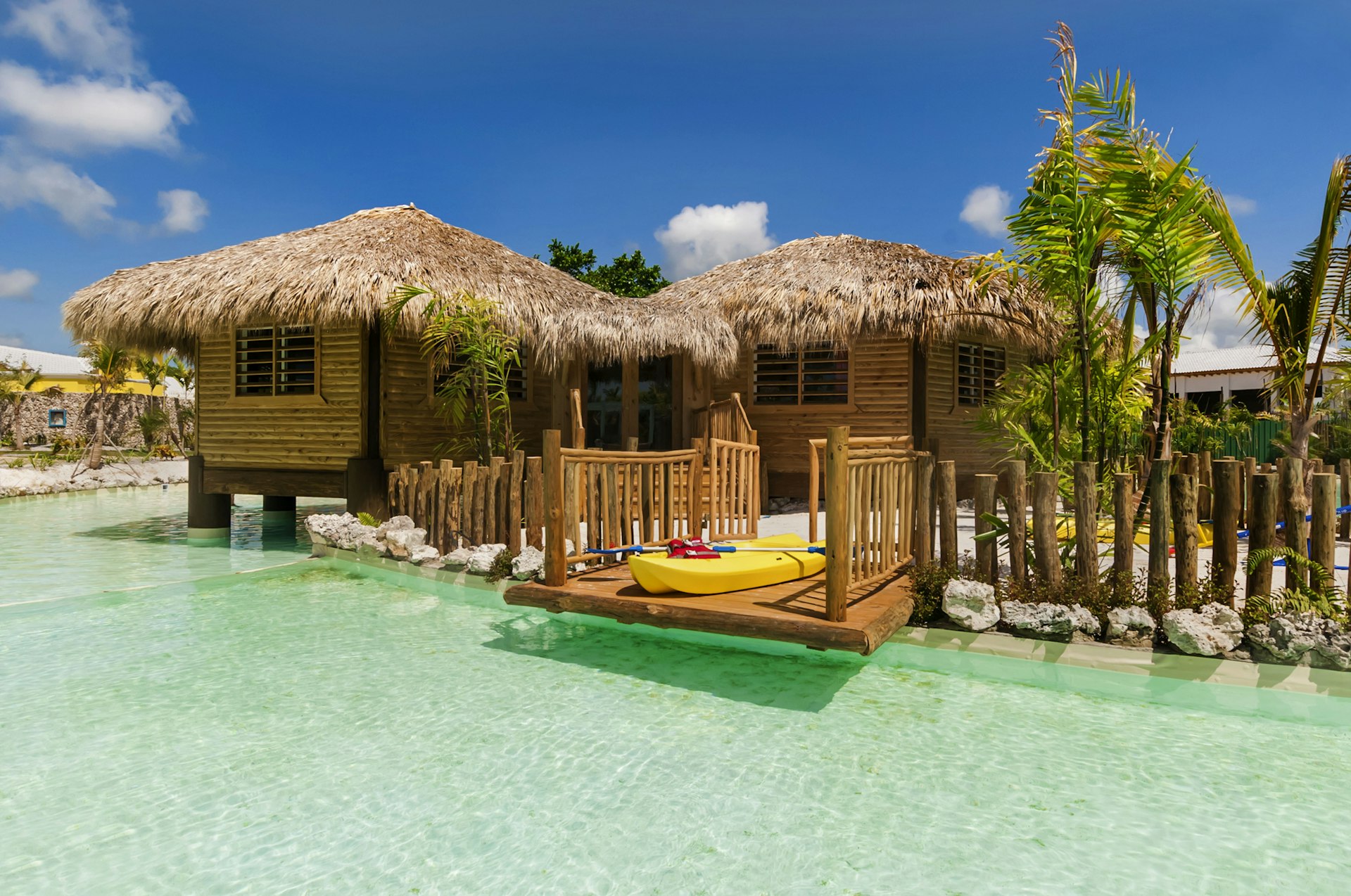 Two wooden houses with straw roofs overlook a body of clear blue water. A kayak is visible.