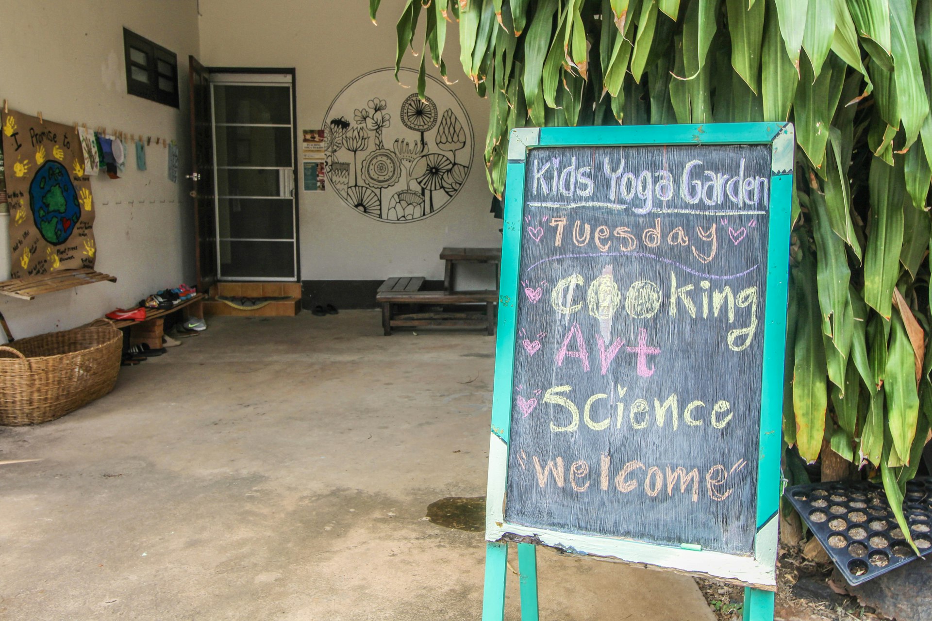 A freestanding black chalkboard sign stands outside a building. The sign reads: "Kids Yoga Garden. Tuesday: cooking, art, science. Welcome".