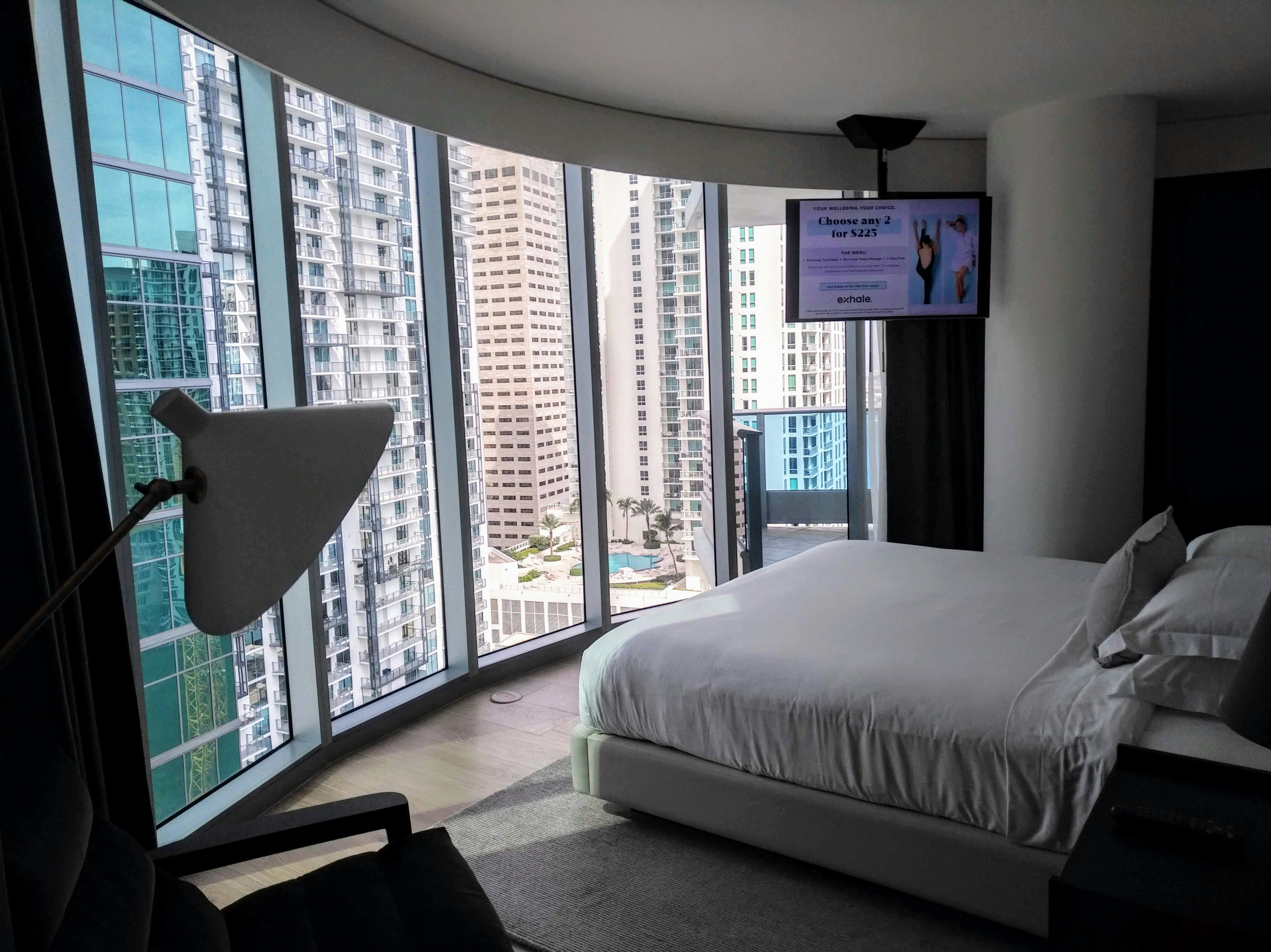A room at the Kimpton Epic hotel in Miami has a curved class wall made up of five narrow rectangular panes overlooking a sea of skyscrapers outside. Inside, a bed faces the curved window wall, made up with simple white linens on a grey woven rug. A tv is mounted to the ceiling and advertises wellness packages. A lamp and chair are just visible in the foreground.