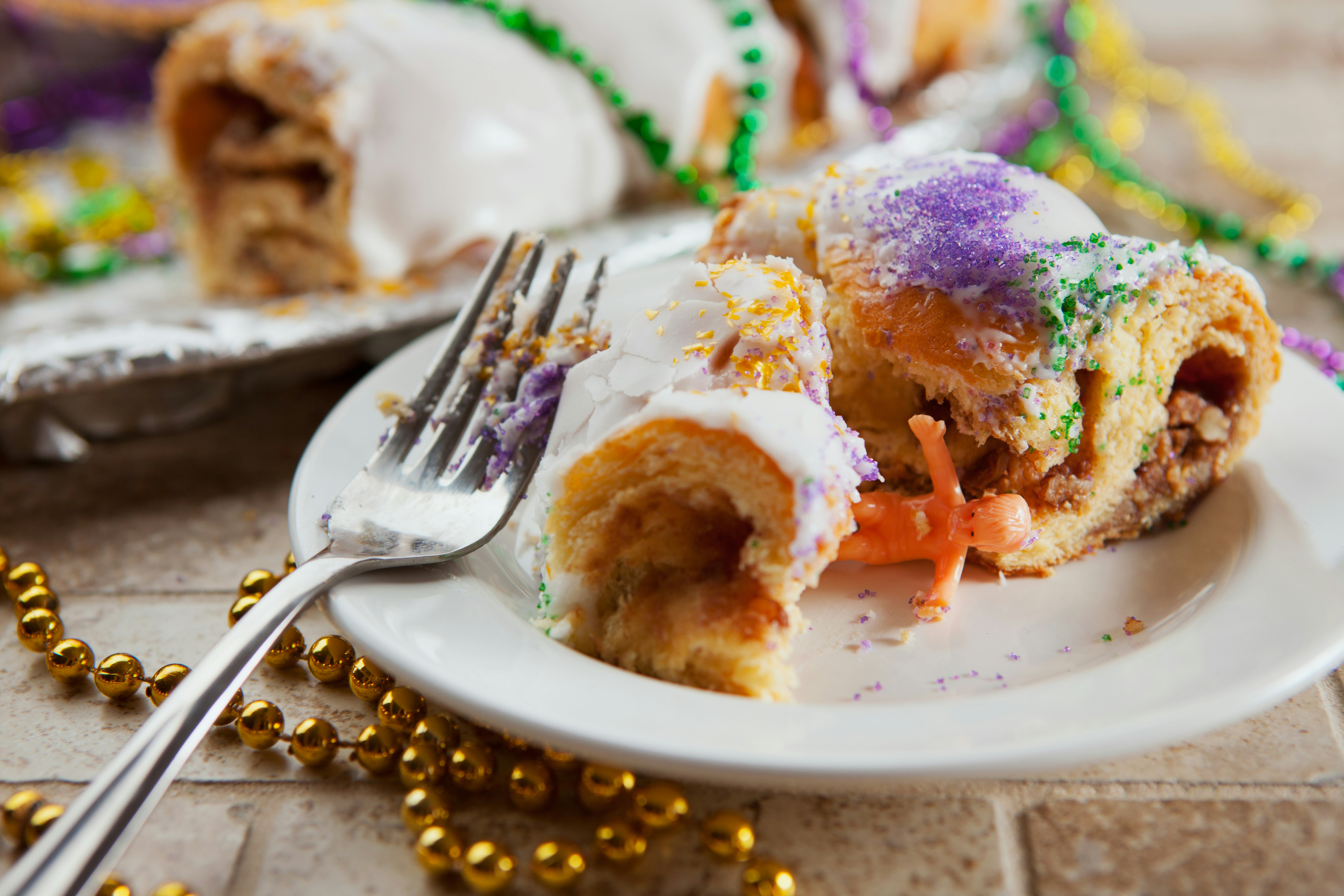 A fork covered in purple frosting rests on the side of a plate that has a slice of cake covered in white frosting and pink, yellow and green sprinkles. There is a plastic baby in between the two pieces of cake.