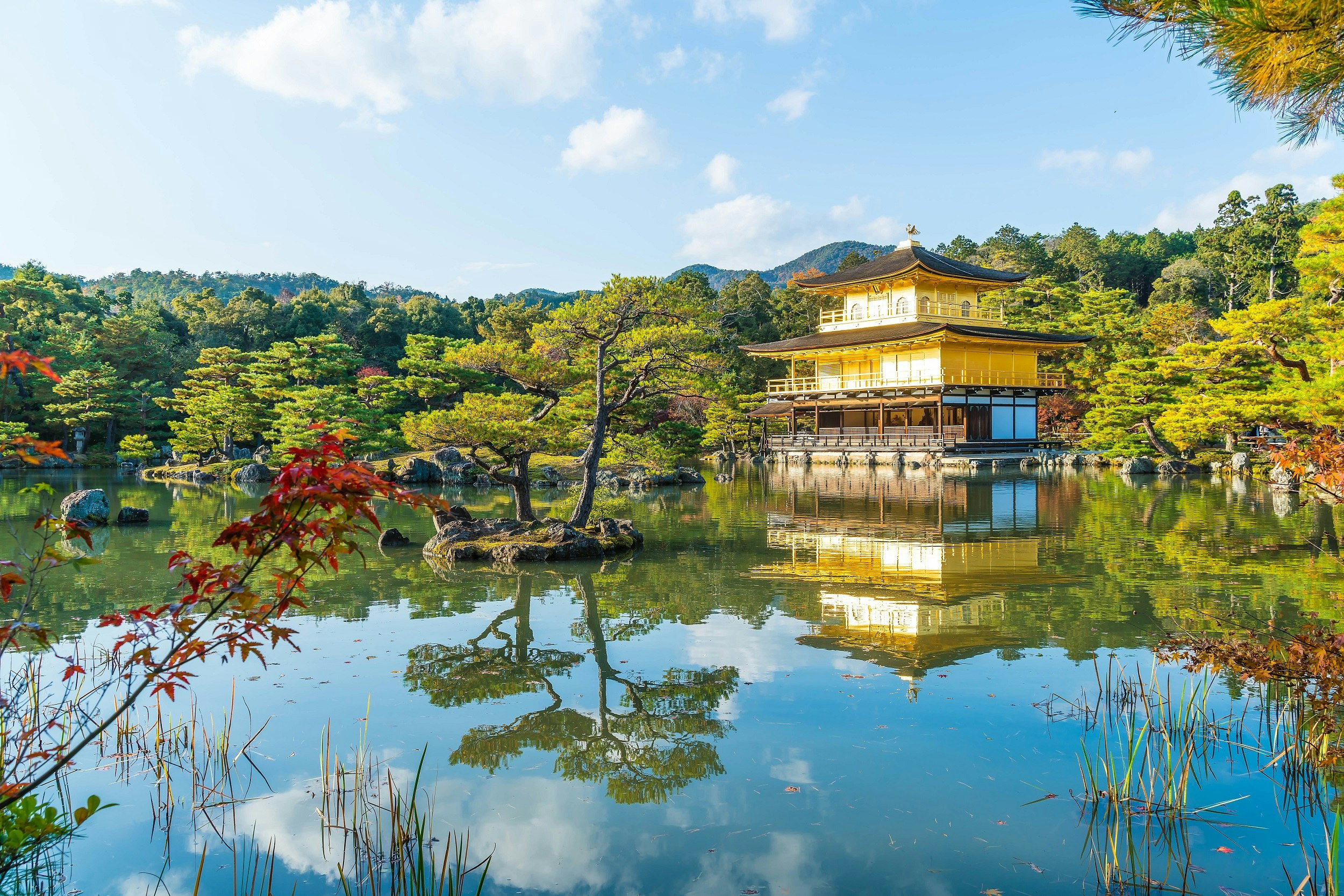 Beautiful Architecture at Kinkaku-ji (The Golden Pavilion) reflected in a lake in Kyoto, Japan; bordering the lake is a dense forest, with blue skies above.