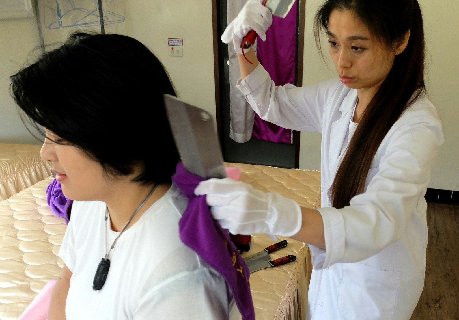 Hsiao Mei-Fang gives a knife massage to one of her students, holding one of the large knives near her shoulder and the other in mid-air. She wears a white doctor's coat with matching white gloves.