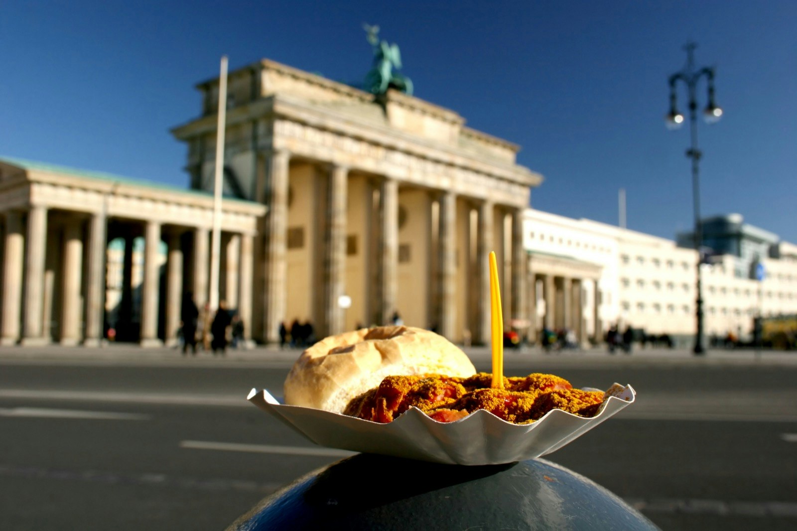 Person balances a currywurst on their knee with the Brandenburg Gate in the background