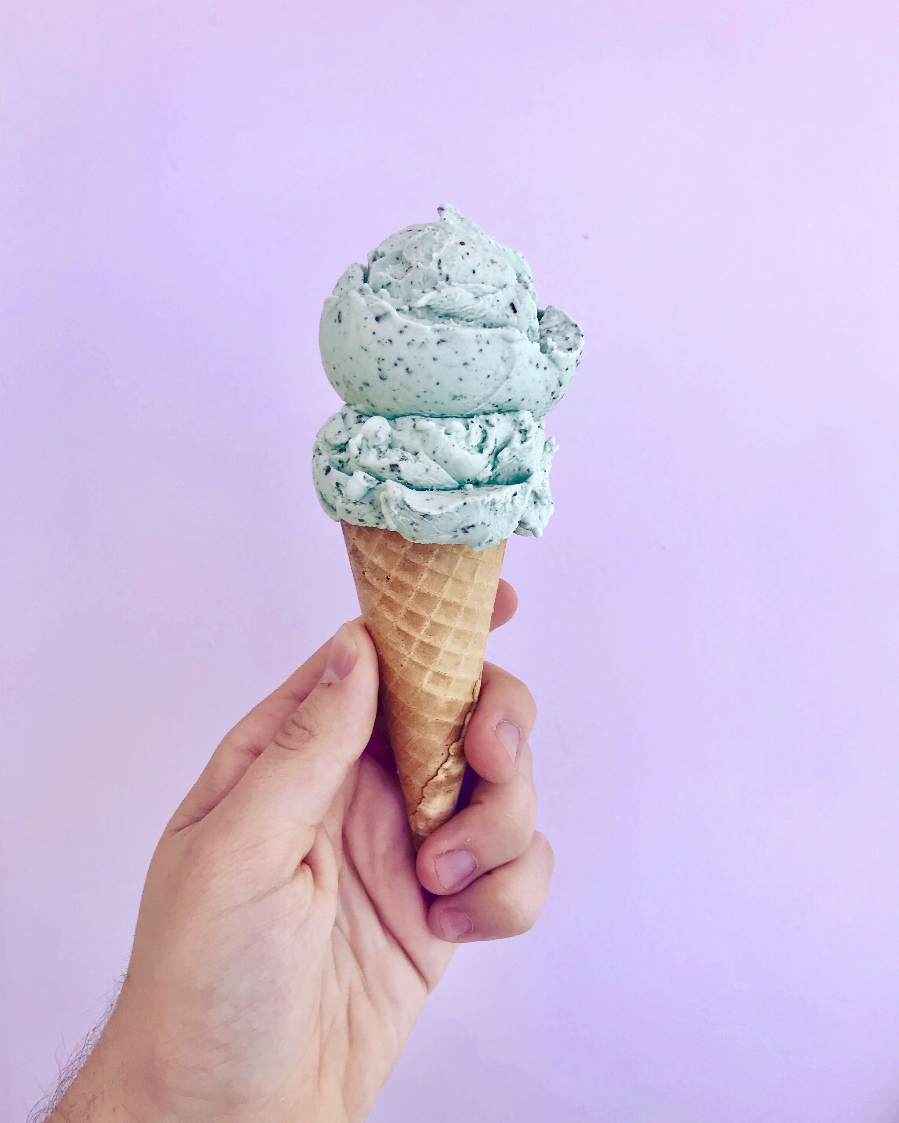 A person holds up a cone with two scoops light green ice cream with flecks of cookie crumbs. 