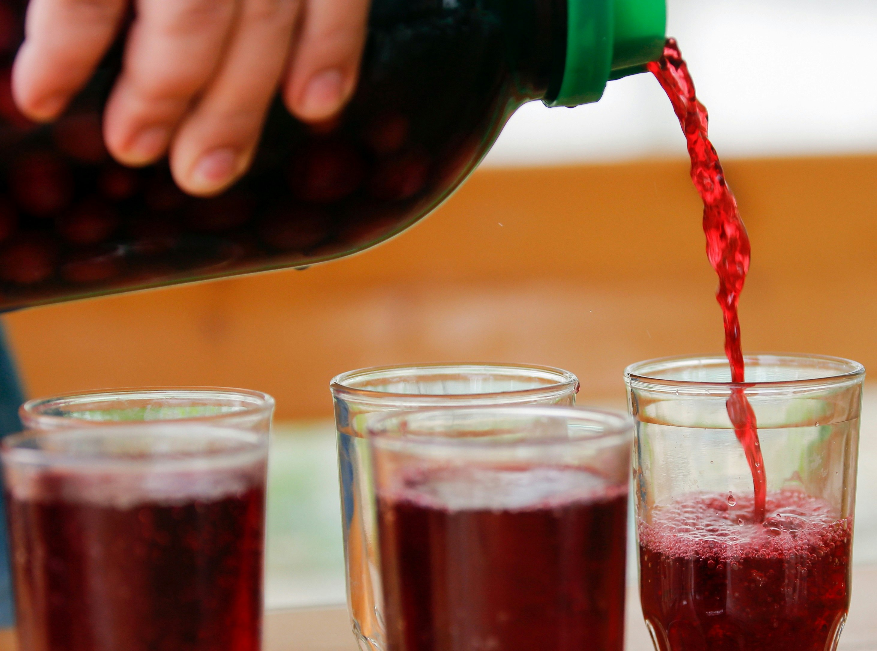 Dark red juice is poured from a bottle into five glasses.