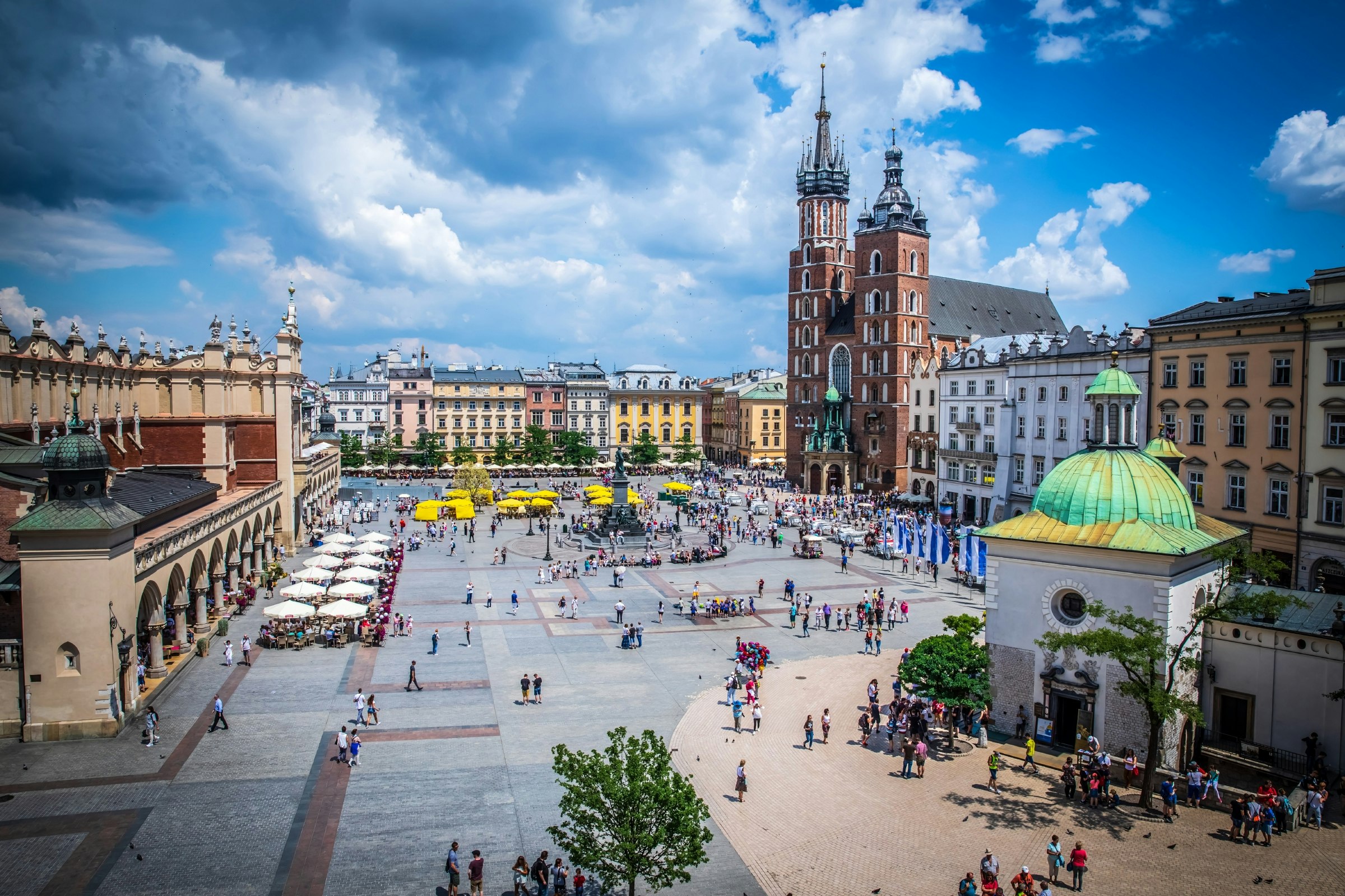 An aerial view of the main square in the Polish city of Kraków. People wander around the large open space, which is flanked by buildings and has a small market place operating in it.