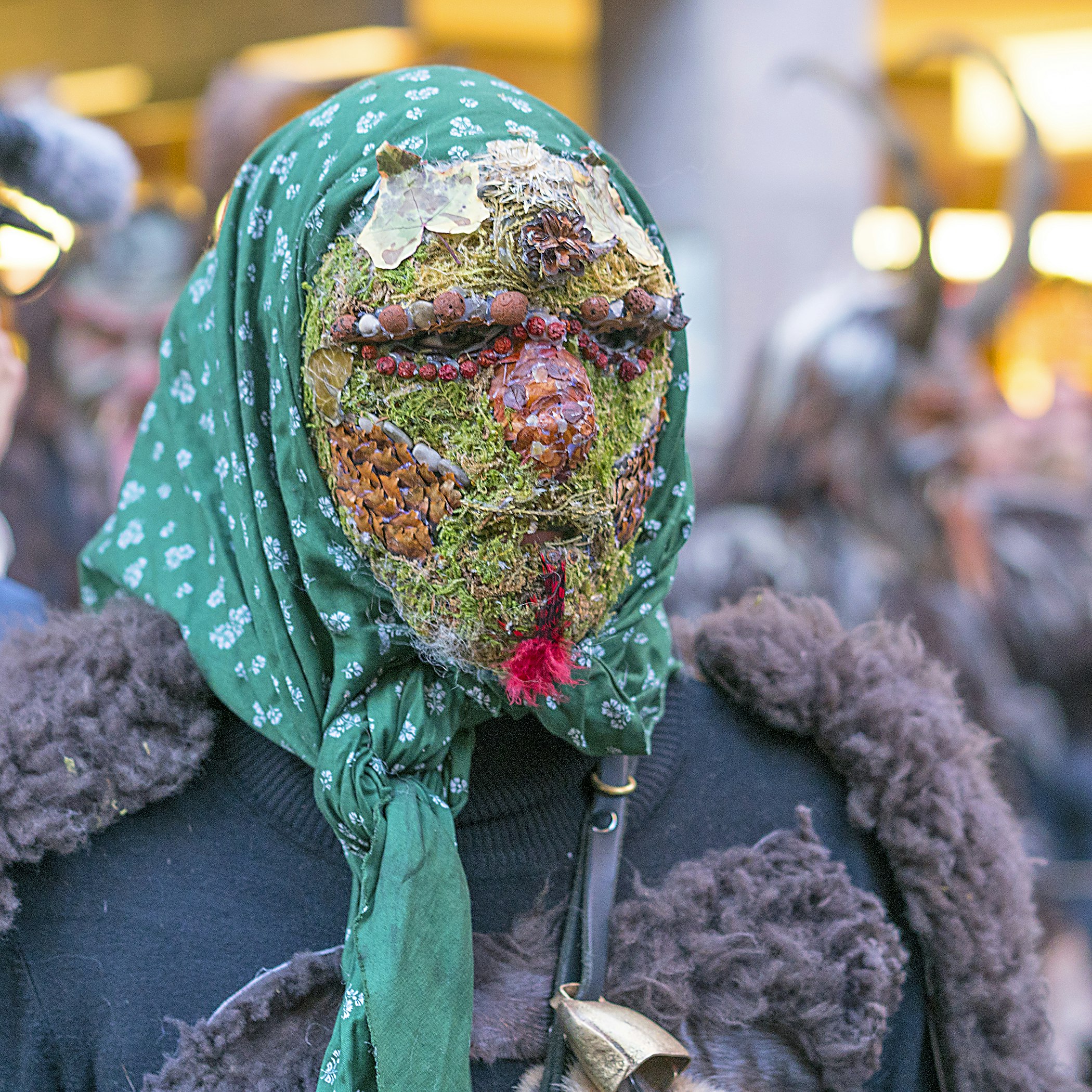 Head and shoulders shot of a person wearing a mask made up of grass, rocks of varying colors and other pieces of shrubbery. The person is also wearing a green and white head scarf, a leather strap with a small cow bell attached and a back sweater with fake fur patches.   