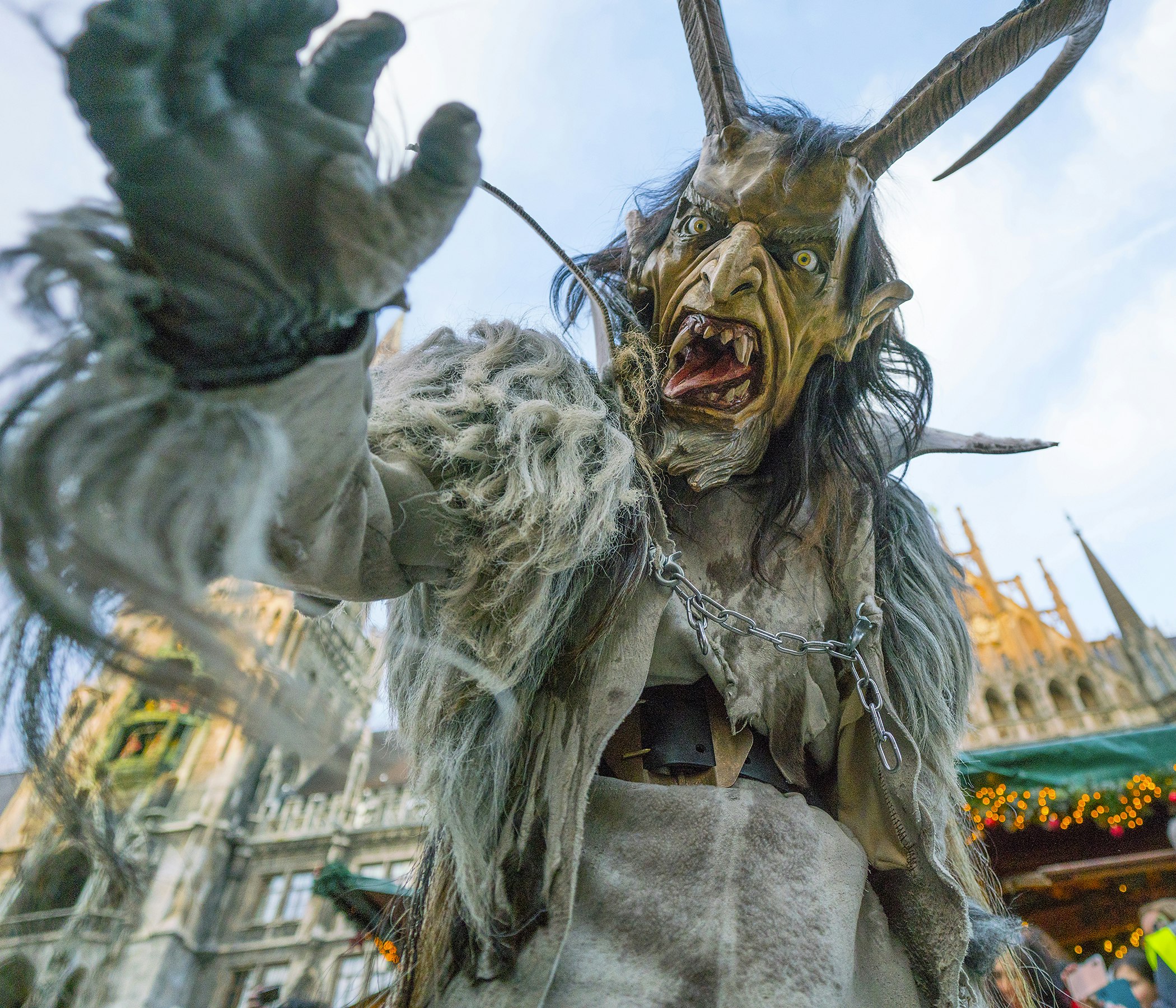 A person dressed as Krampus, the Christmas demon, reaches for the camera while exposing a large tongue and pointy teeth. The Krampus costume also features two large horns protruding from the head and a hairy body  