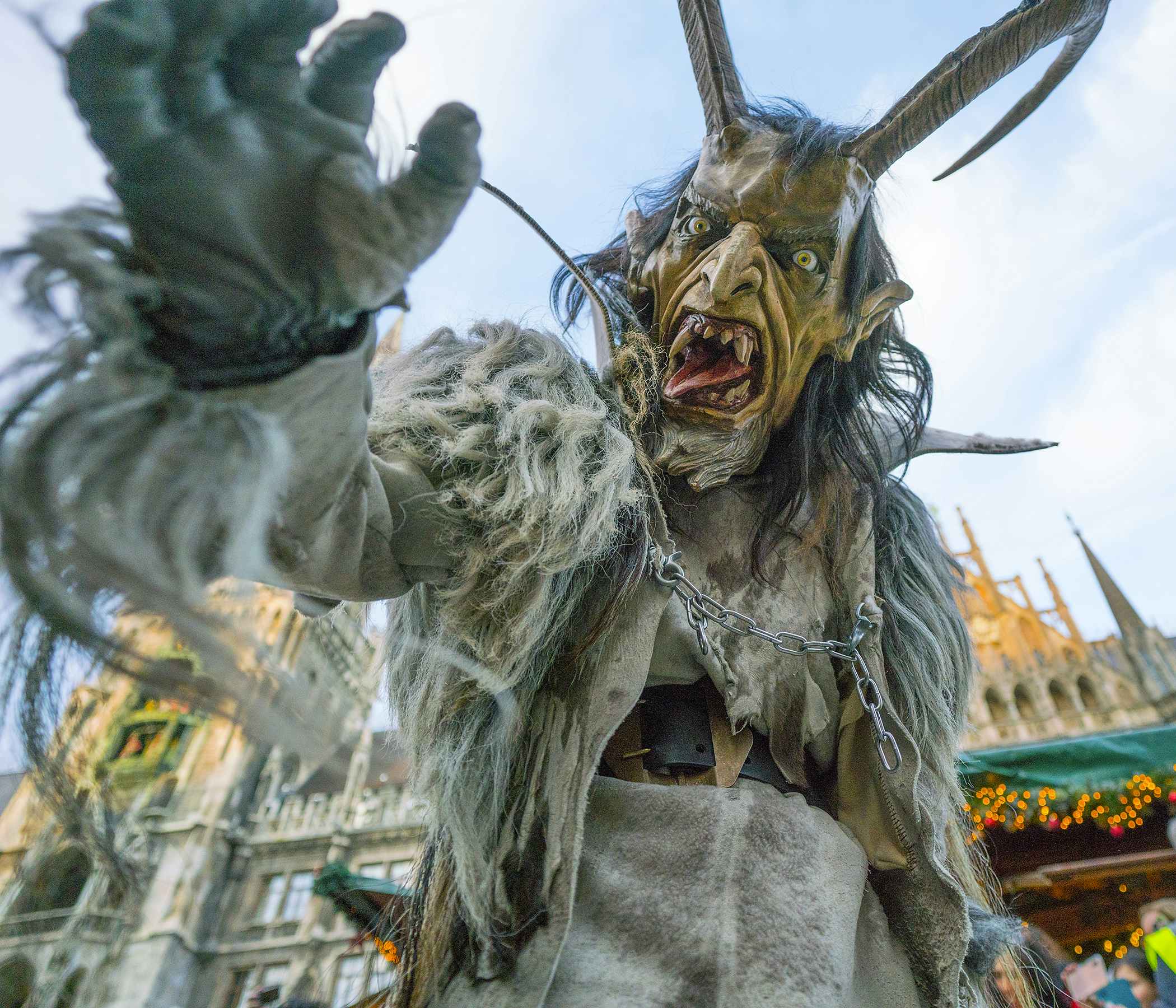 A person dressed as Krampus, the Christmas demon, reaches for the camera while exposing a large tongue and pointy teeth. The Krampus costume also features two large horns protruding from the head and a hairy body