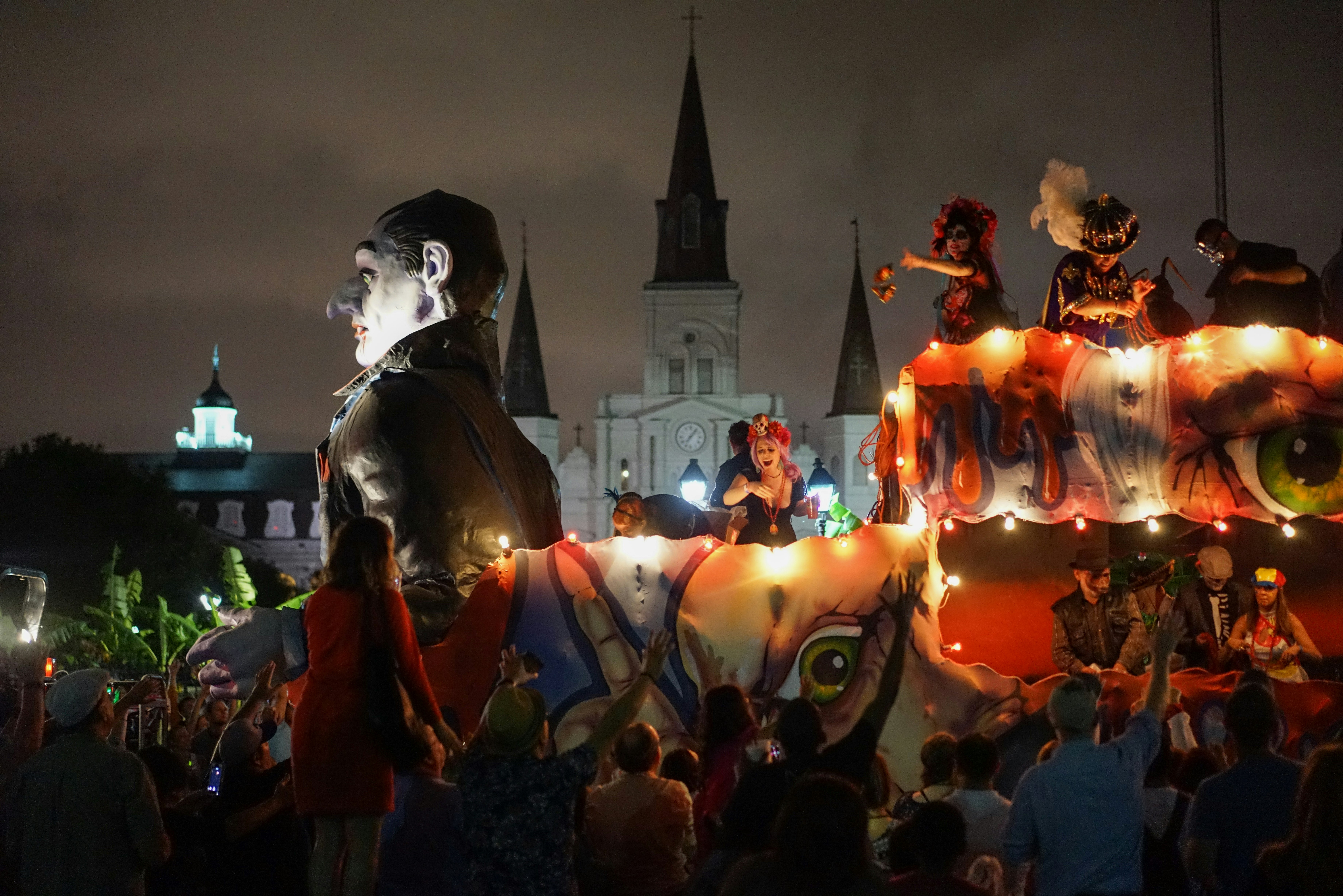A large float with a huge Dracula figure at the front is illuminated by a strong of light bulbs. People dressed in elaborate costumes throw items off the float to people on the street. 
