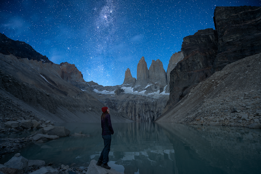 En kvinna står på kanten av en blågrön sjö omgiven av bergstoppar.  Stjärnorna kan ses tydligt på natthimlen.