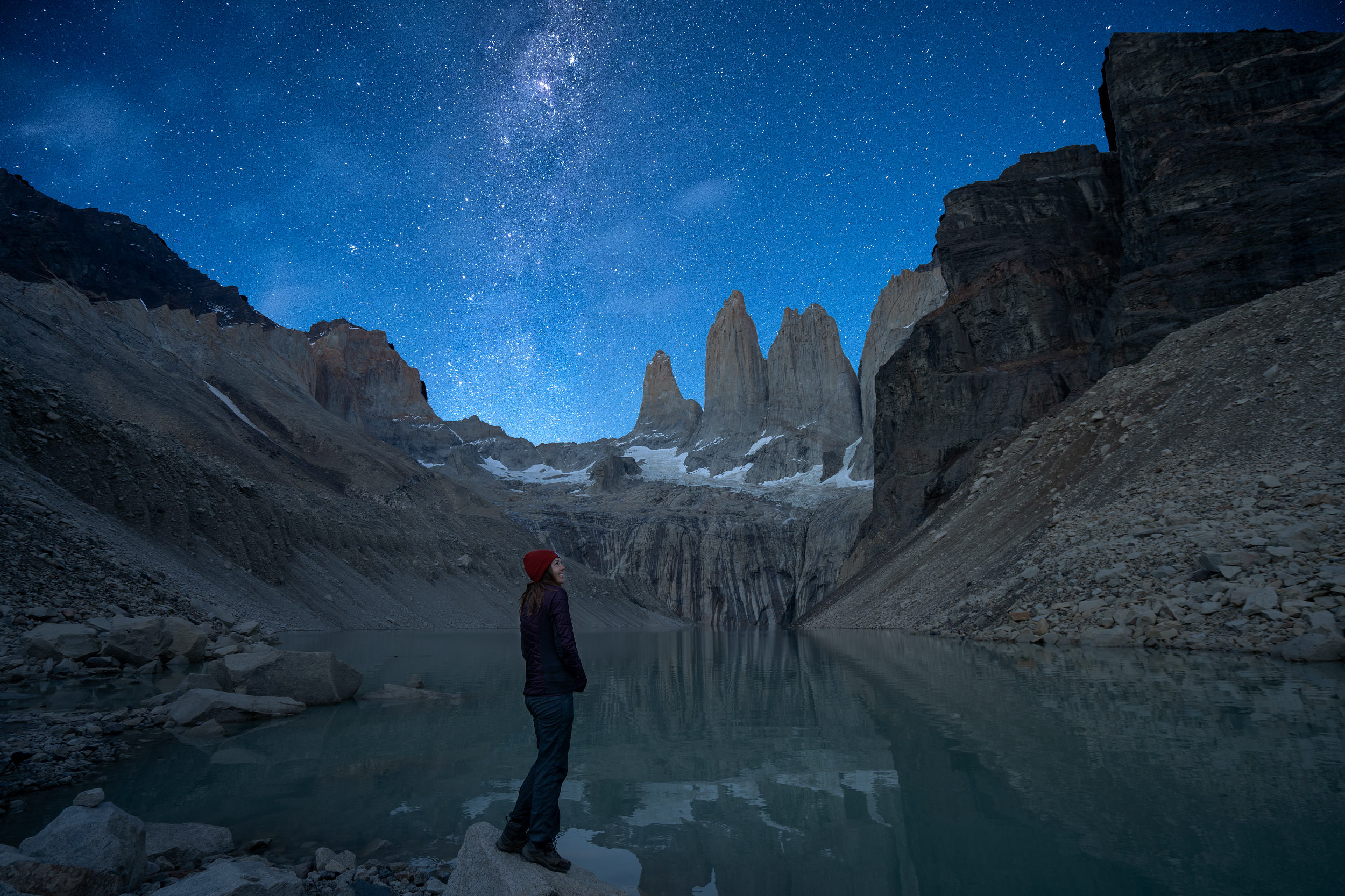 A woman stands on the edge of a blue-green lake surrounded by mountain peaks. The stars can be seen clearly in the night sky.