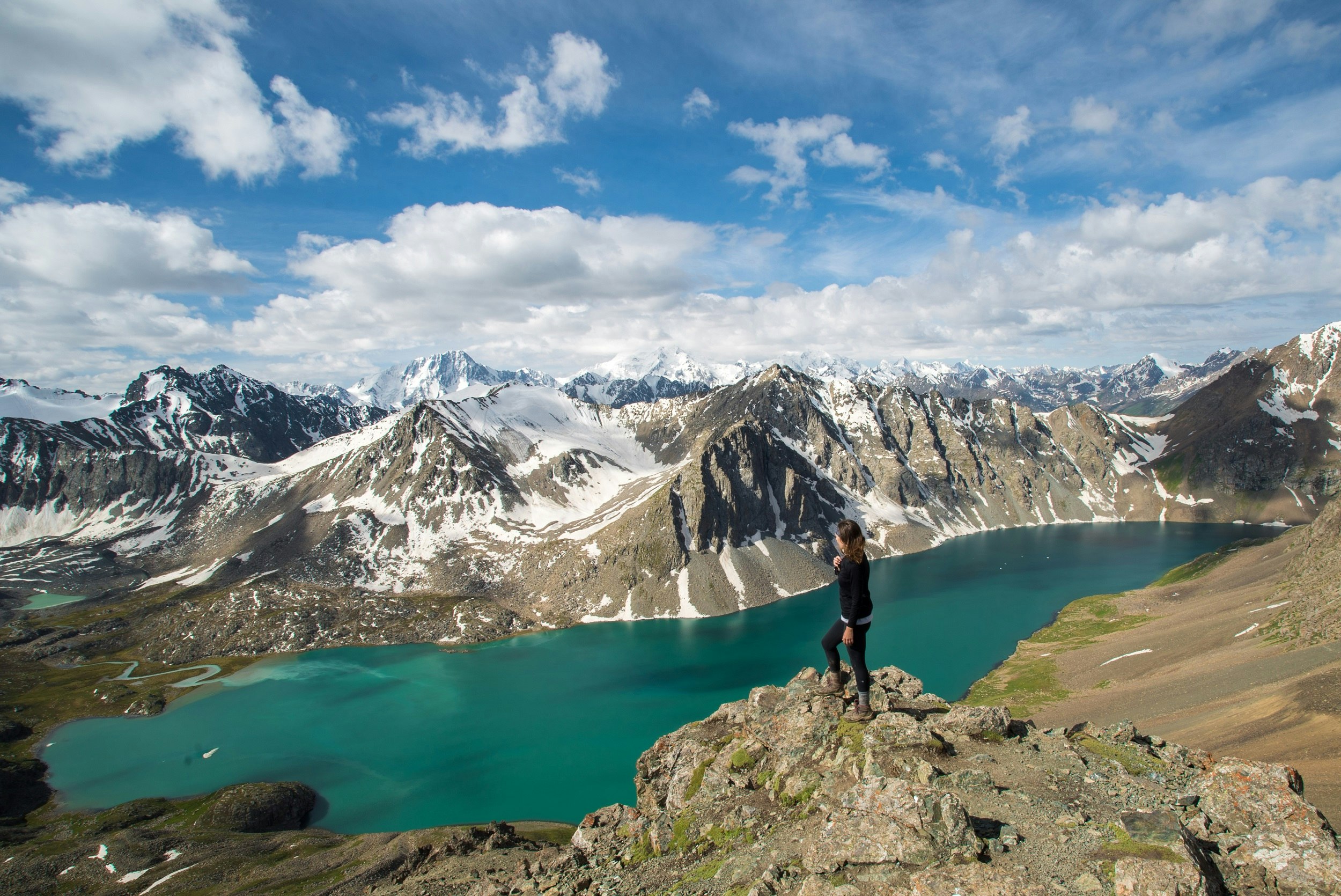 A solo woman stands on a mountaintop surrounded by snowy peaks, with a blue-green lake below.