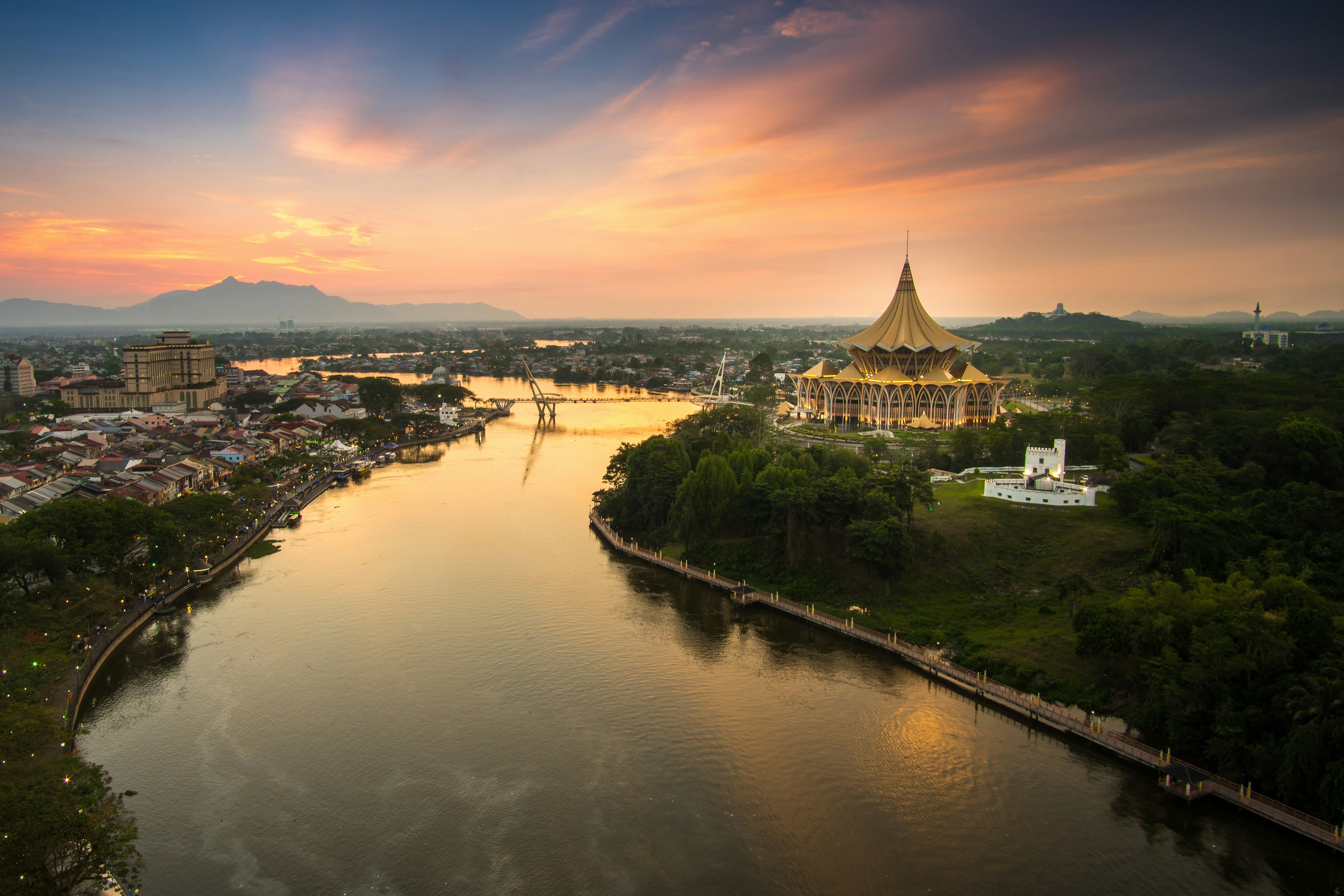 An aerial view of the city of Kuching in Sarawak, Borneo. The image shows the city's river winding away into the distance, passing large buildings and grand temples. In the distance, a mountain range is visible.