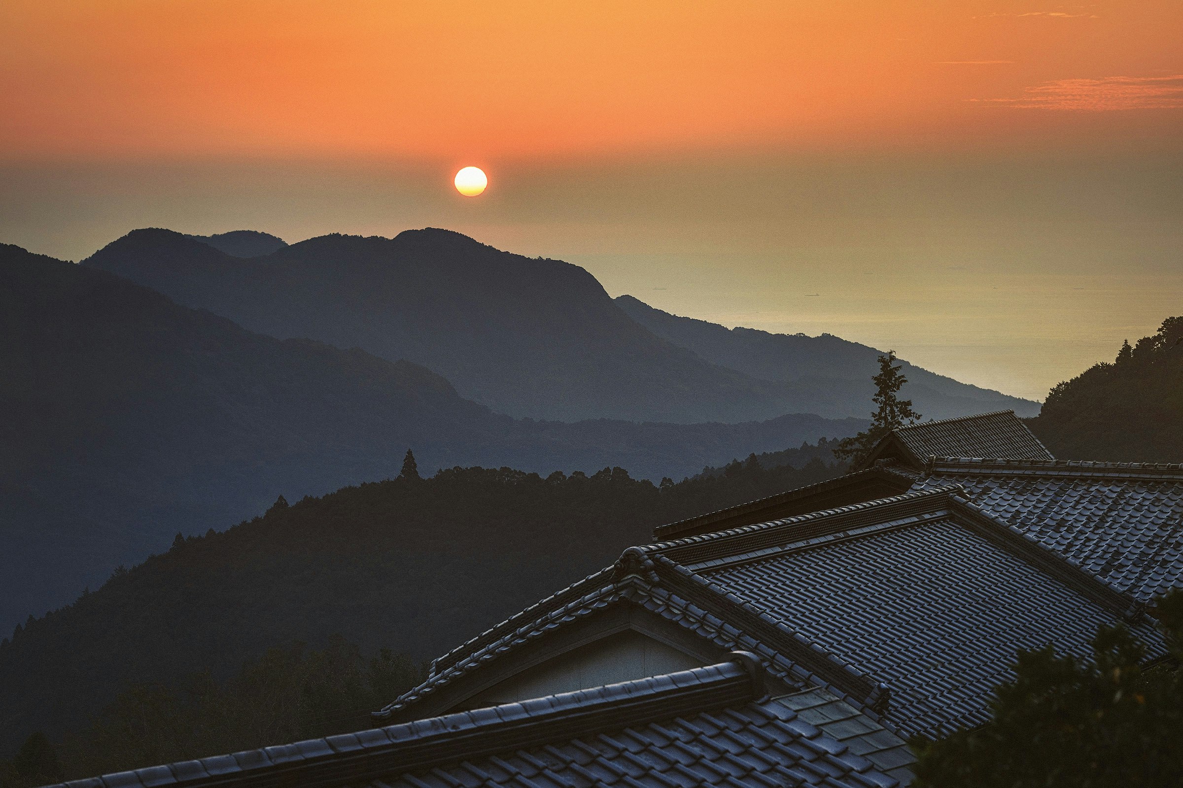 The sun rises over darkened mountains and the rooftops of Nachi Taisha