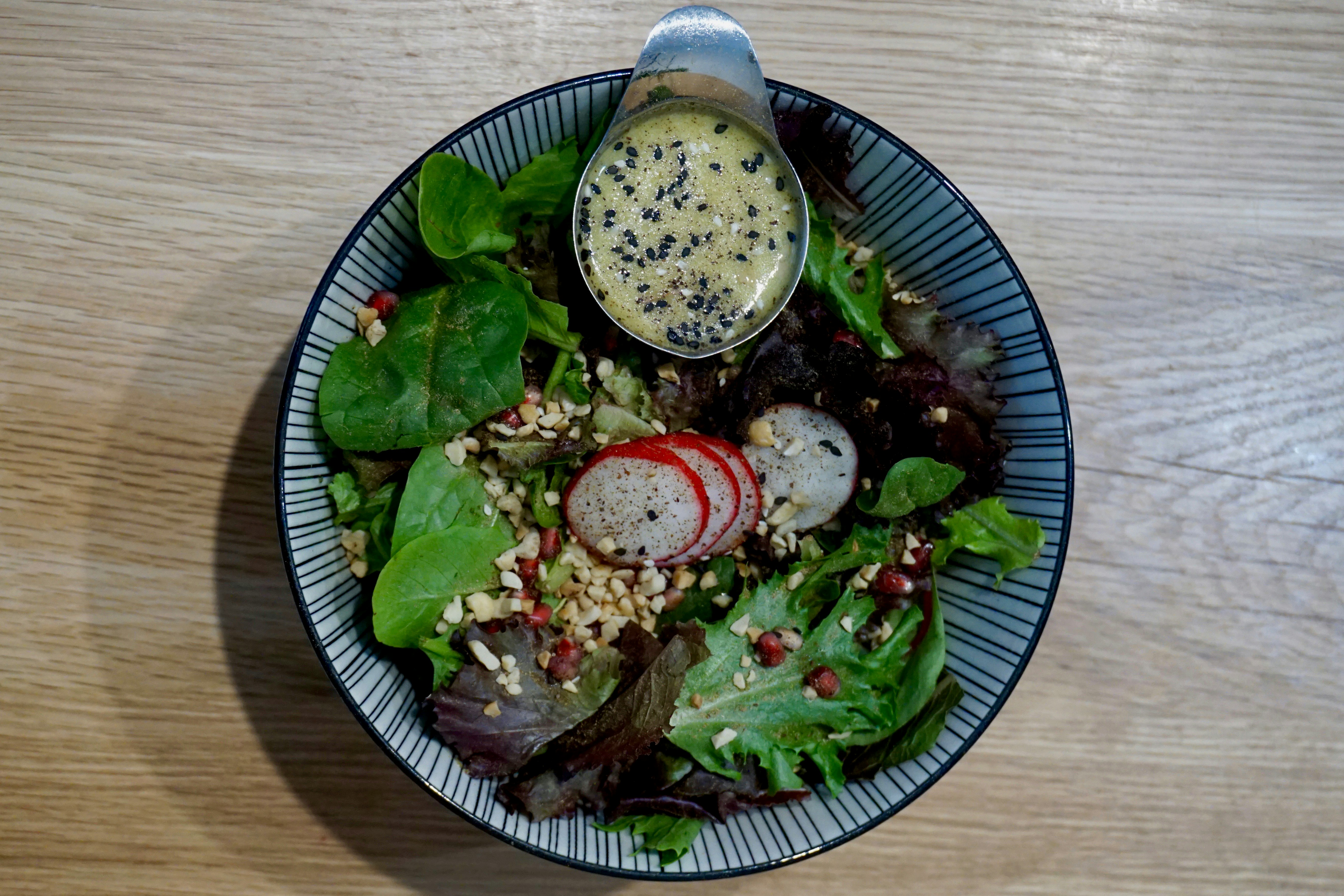 A stripy ceramic bowl sitting on a blond wood table. The bowl is filled with leaves and colourful salad ingredients and there's a stainless steel ramekin filled with dressing on the side.