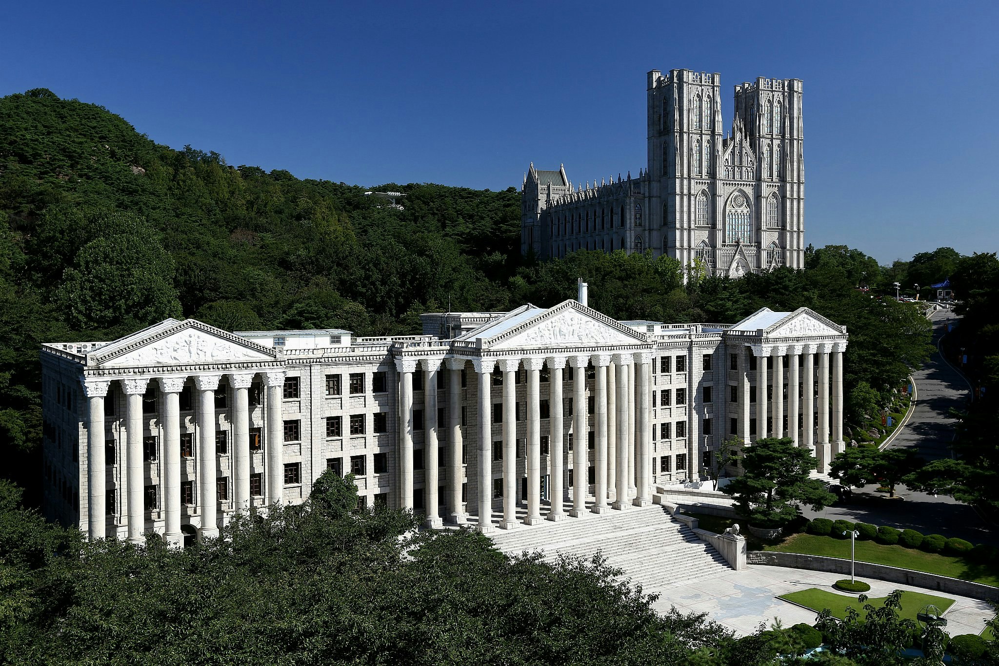 A pair of buildings on the Kyung Hee University campus in Seoul, South Korea: the white, colonnaded University Administration Building, reminiscent of a Greek temple, in the foreground, with the ornate Grand Peace Hall behind, resembling a cathedral. 