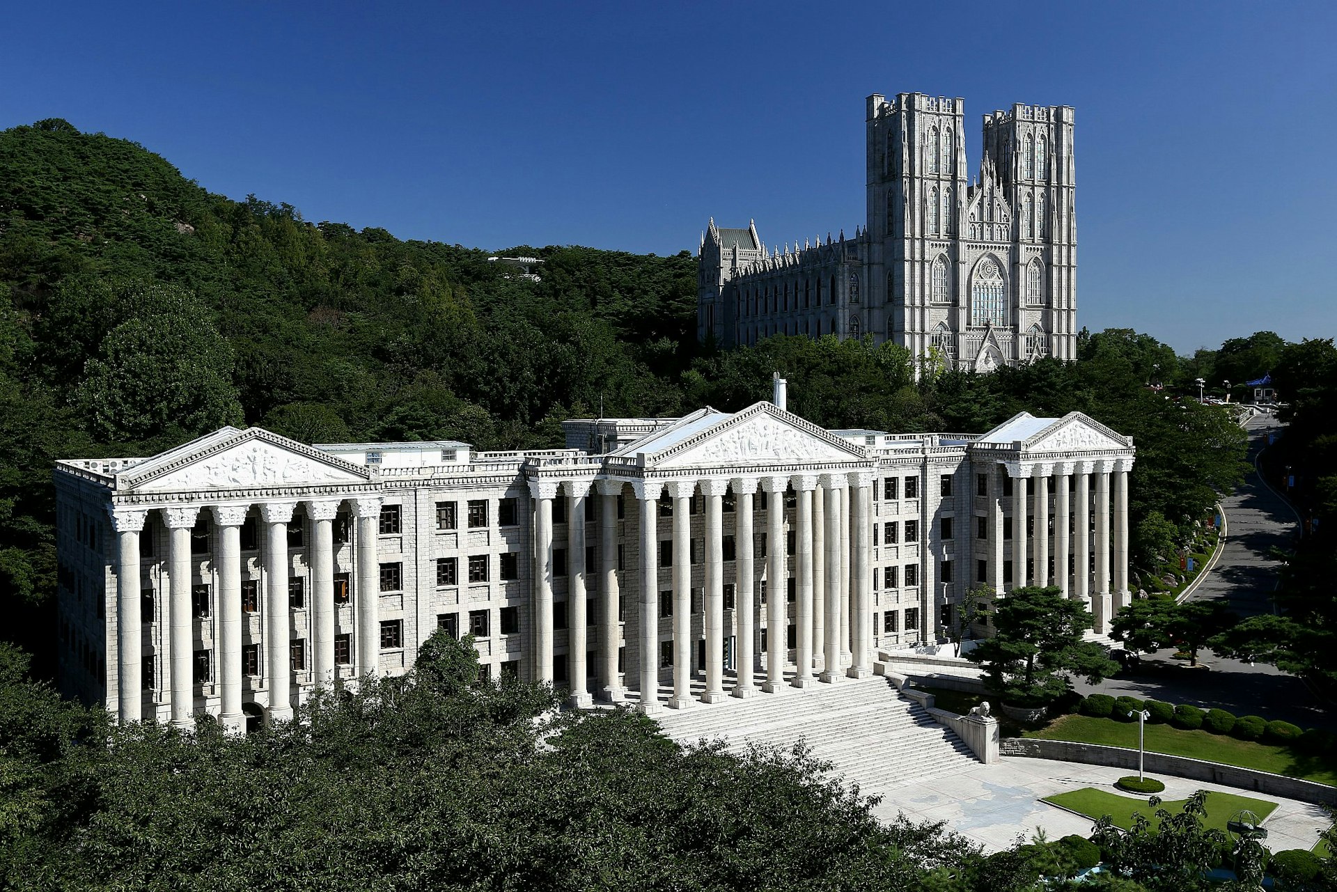 A pair of buildings on the Kyung Hee University campus in Seoul, South Korea: the white, colonnaded University Administration Building, reminiscent of a Greek temple, in the foreground, with the ornate Grand Peace Hall behind, resembling a cathedral. 