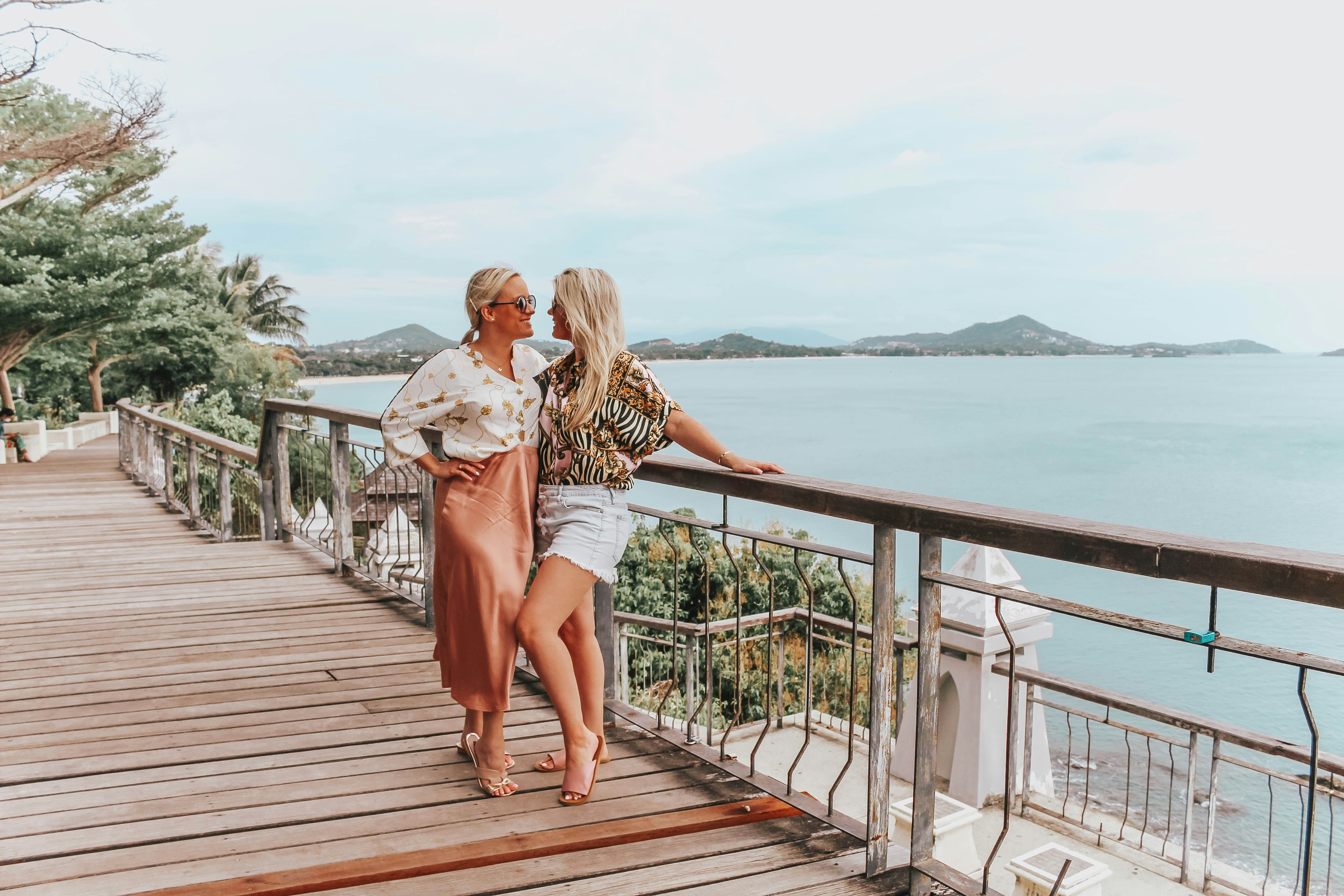 Whitney and Megan embracing on a tree-lined, tropical coastal boardwalk on Ko Samui, with the water and other islands visible behind.