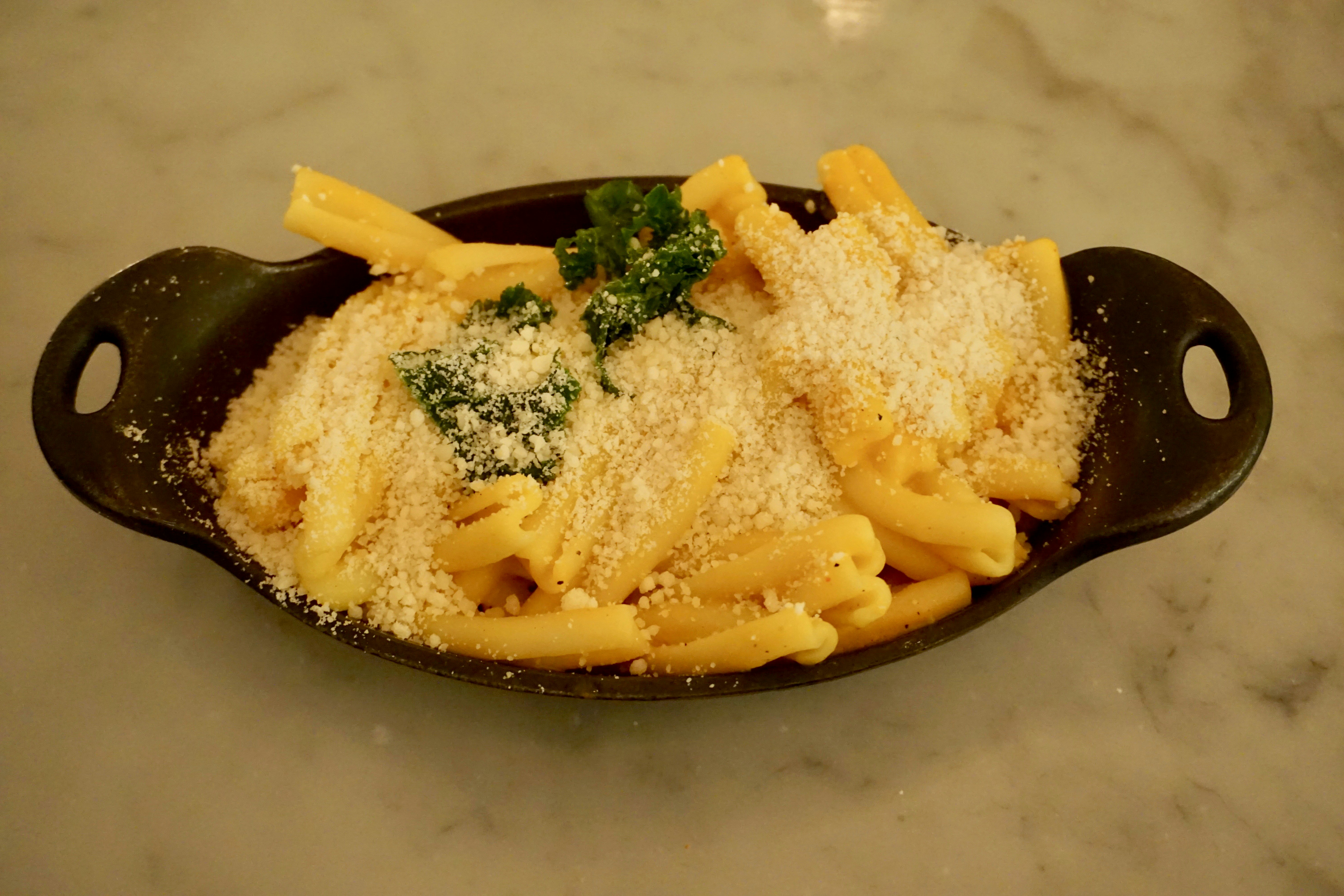 A brown oval dish is filled with mac n cheese; the pasta is sprinked with vegan parmesan and is garnished by some sprigs of kale.