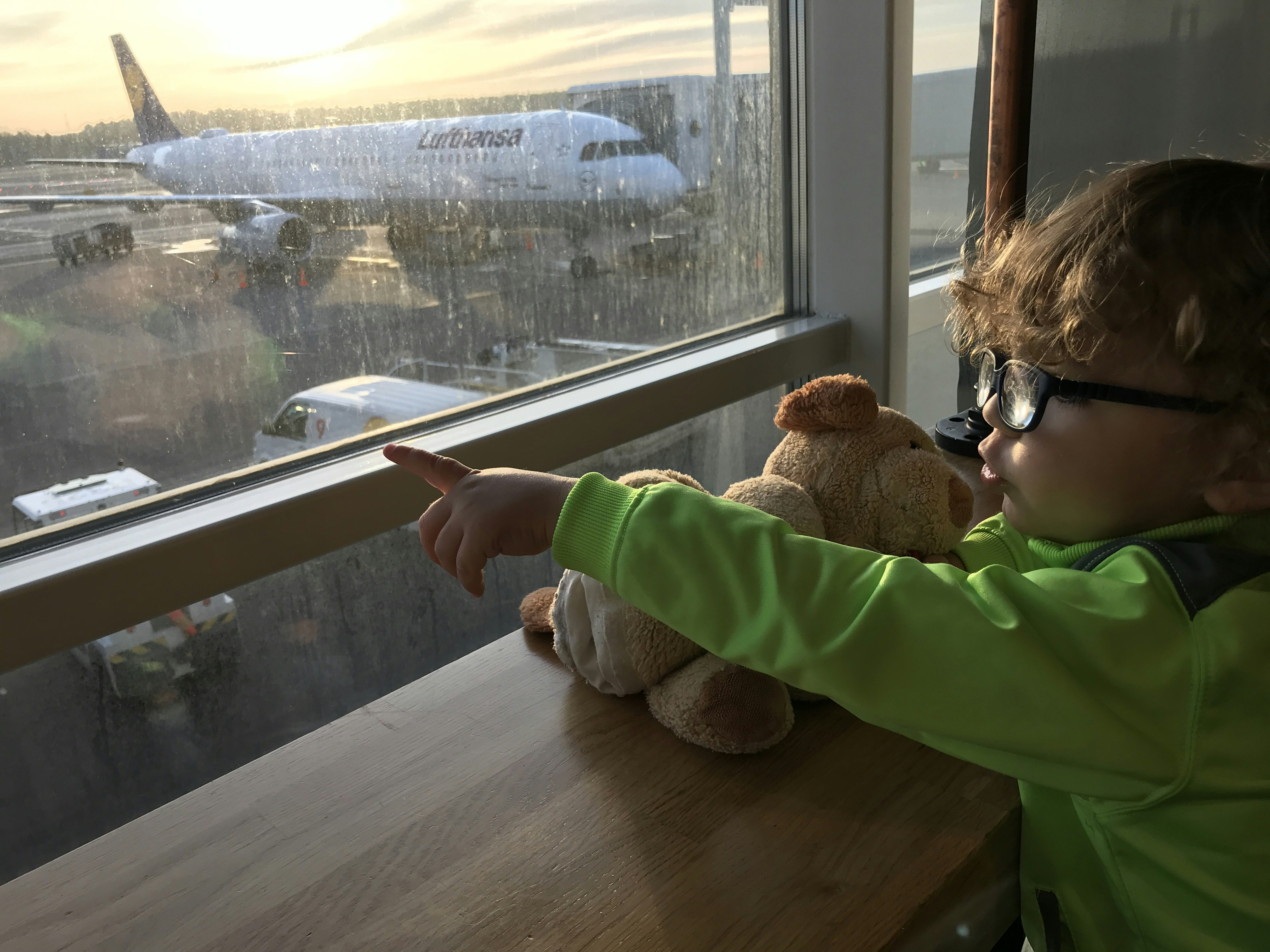 A small child is looking out of an airport window and pointing at something outside.