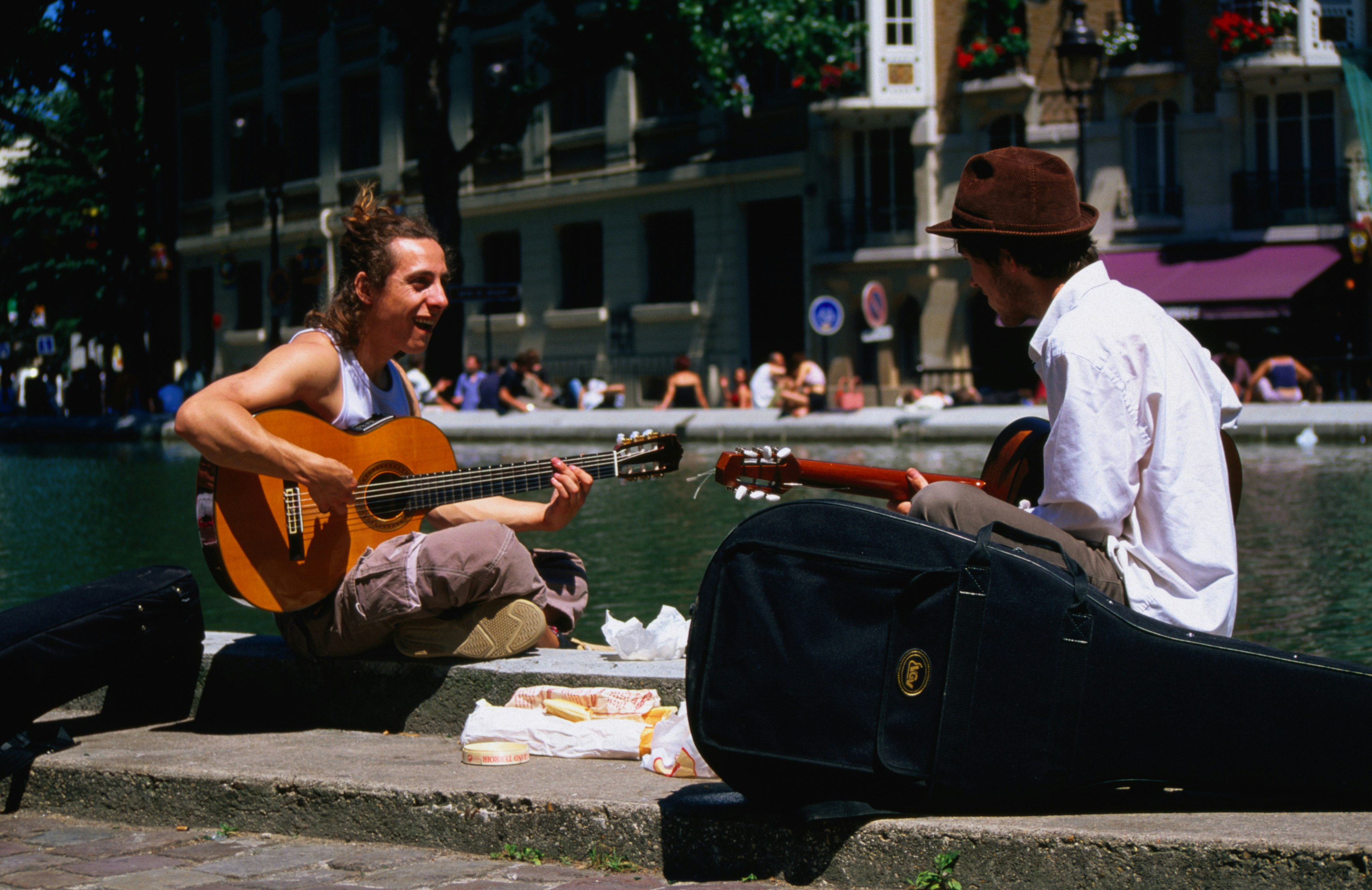 Two men sit on the banks of the canal playing guitars with a picnic of bread, cheese and fruit next to them