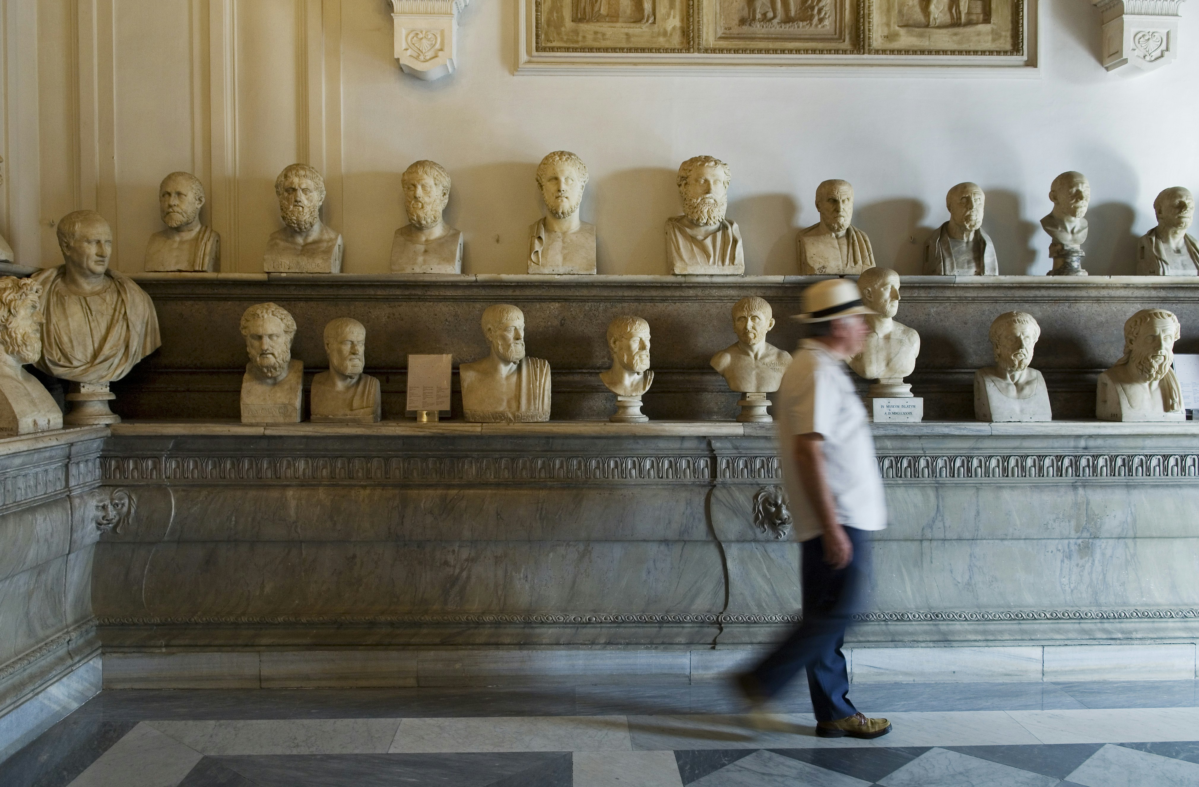 An array of marble busts of various philosophers are sitting on shelves in a museum. A man is walking past.