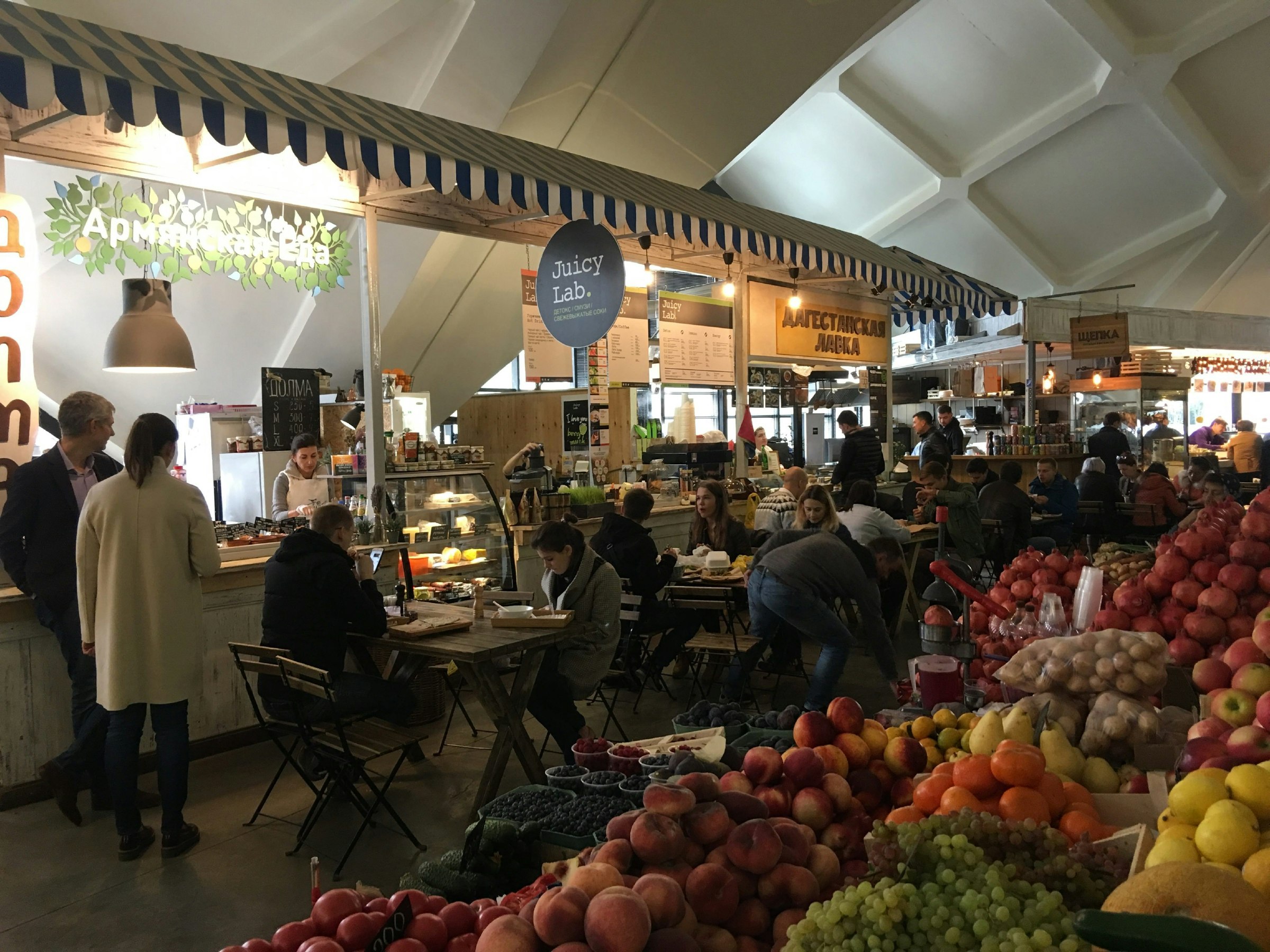 Cafes and food stalls in a covered market. There are piles of fruits and vegetables in the foreground