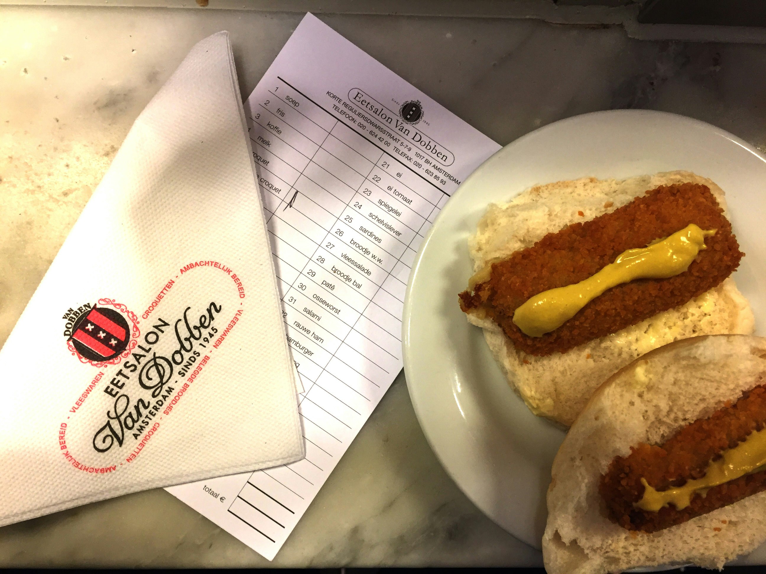 On the right is a bread roll split in half; each slice is topped with a kroket and a line of yellow-brown mustard. On the left is a napkin folded diagonally, displaying the name of the cafe (Eetsalon Van Dobben).