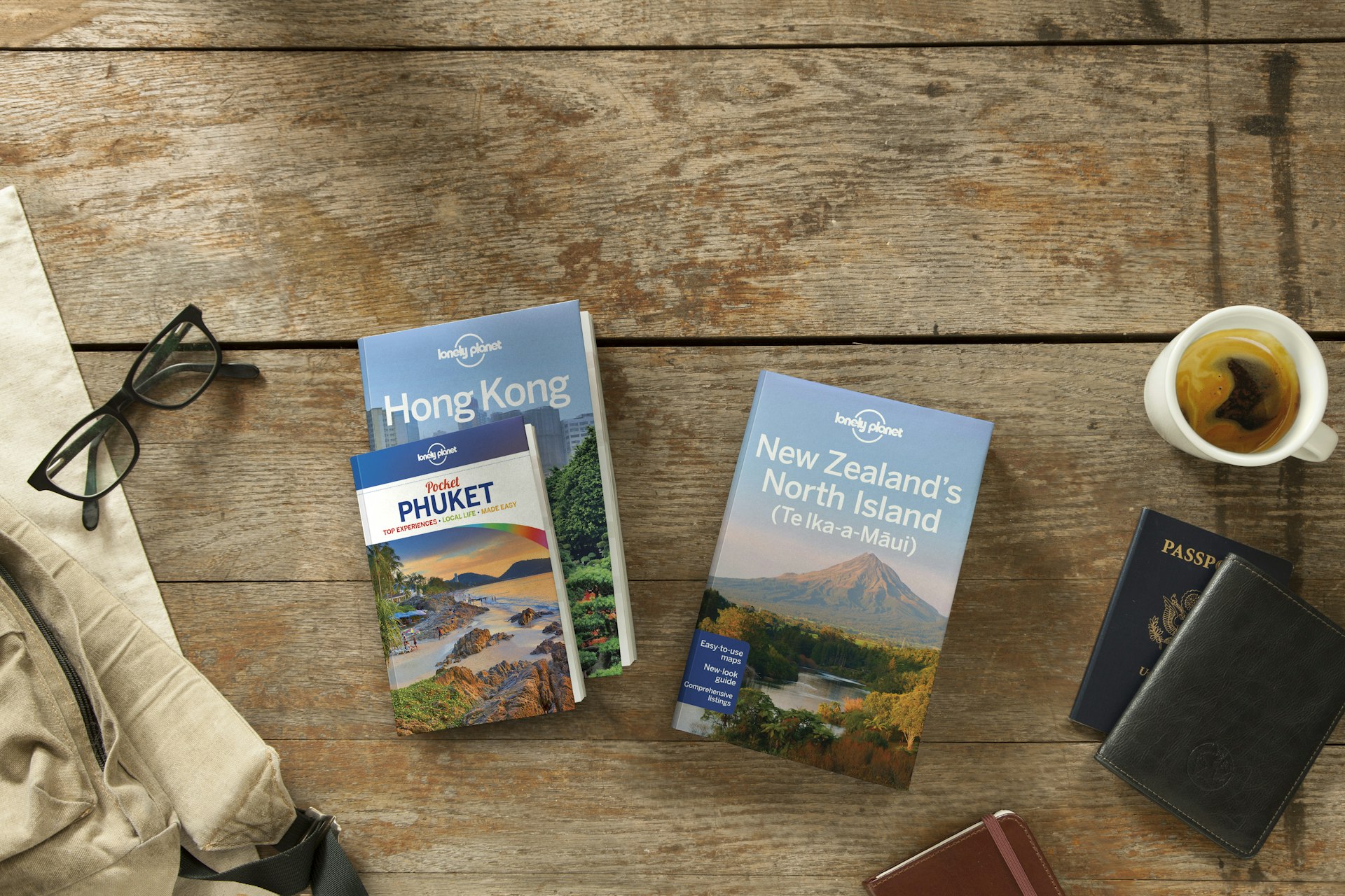 Several Lonely Planet guidebooks sit on a warm, honey-hued wooden table with large planks. One is a guide to Hong Kong, another to Phuket. A pair of black eye glasses sit off to the side. Another is a guide to the north islands of New Zealand, sitting by a blue American passport and a mug of herbal tea.
