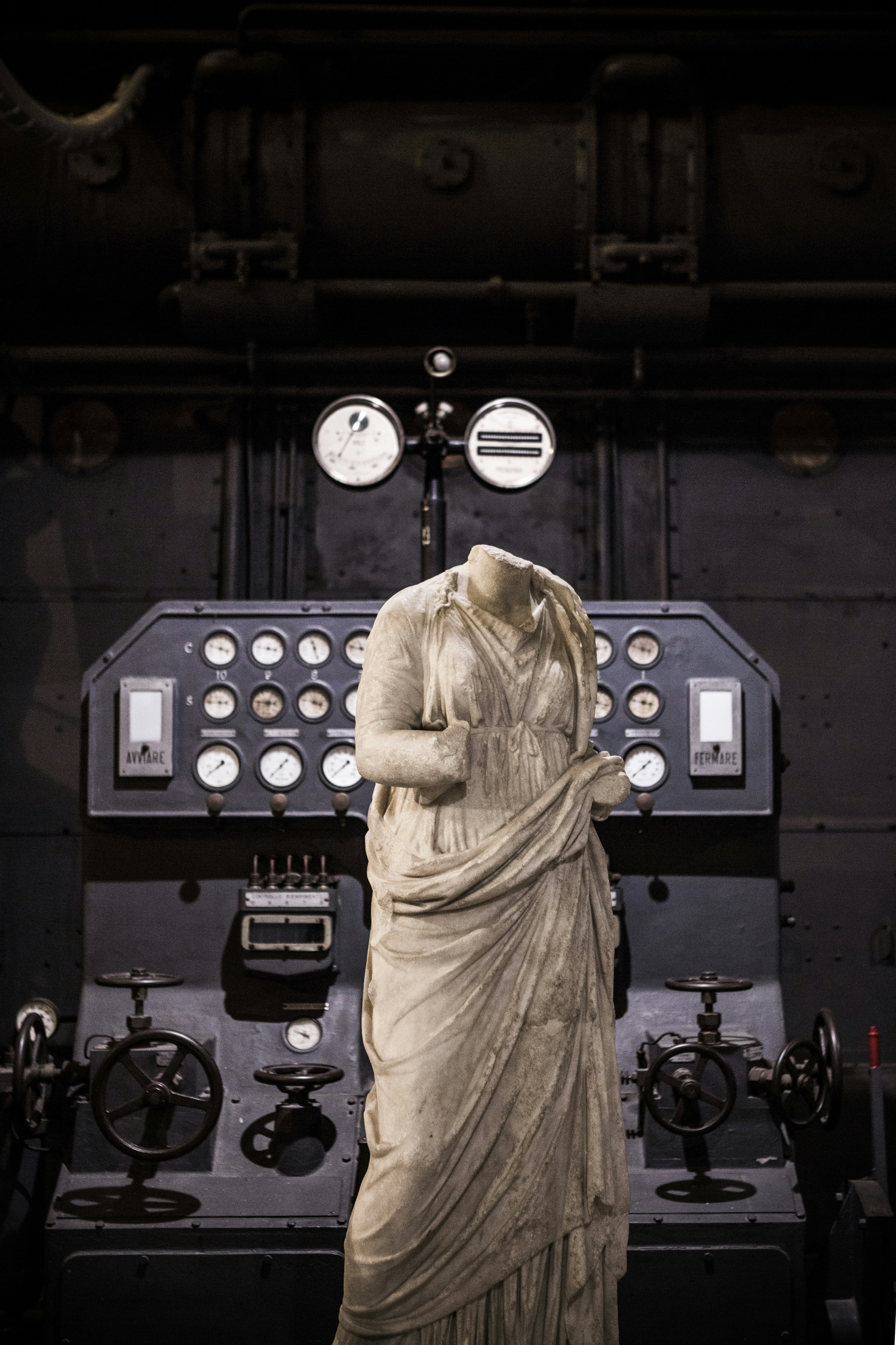 A white, headless, marble statue stands in front of an old black control station that has many dials and gauges.