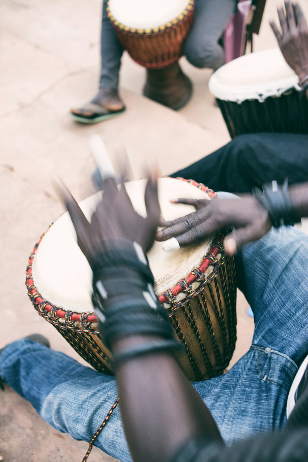 A pair of blurred hands play a djembe that is held between the musician's legs; he's wearing blue jeans.