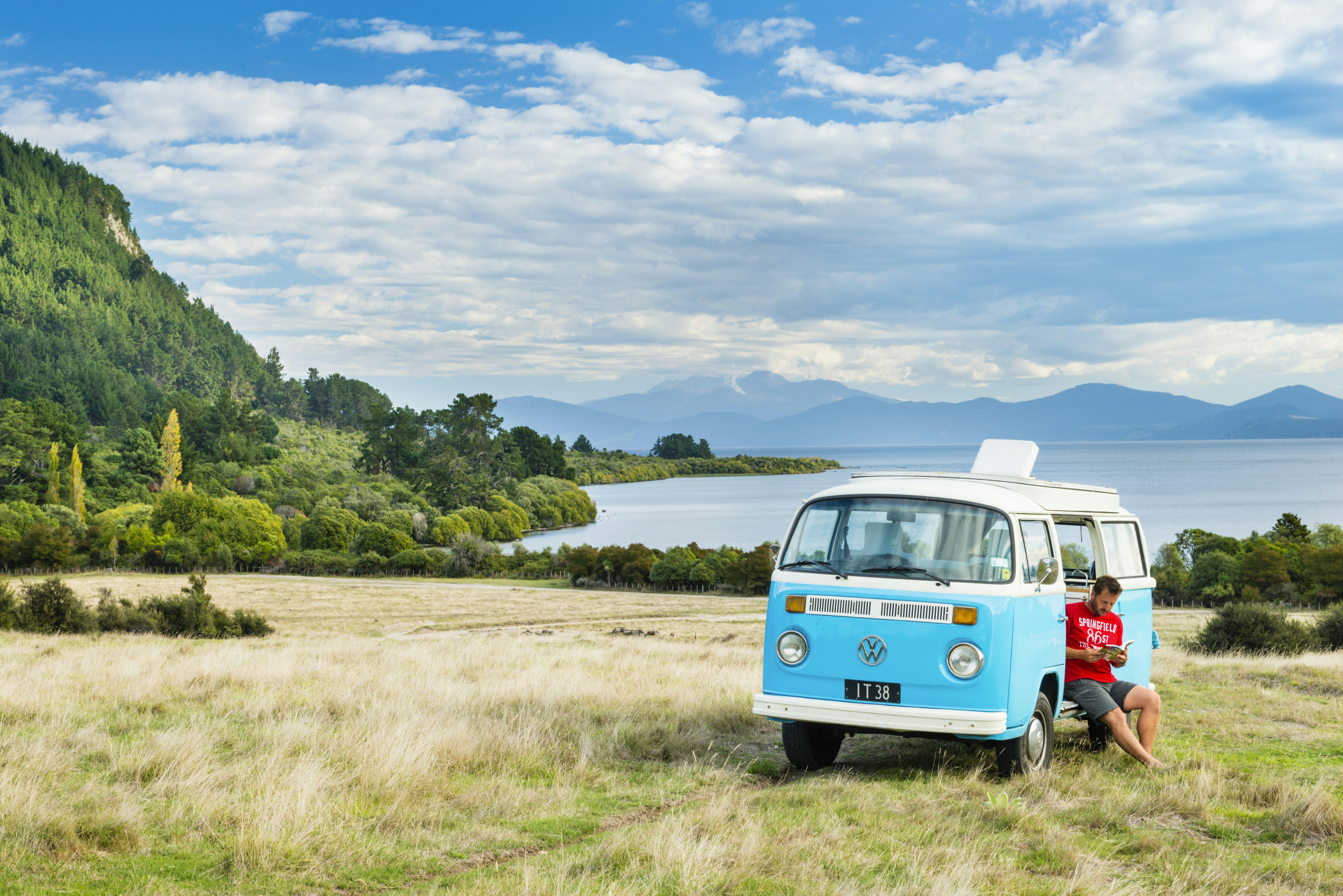 A man in a red T-shirt is sitting in the open door of a bright blue VW campervan reading. The can is parked on grassy terrain beside a beautiful lake lined with green trees.