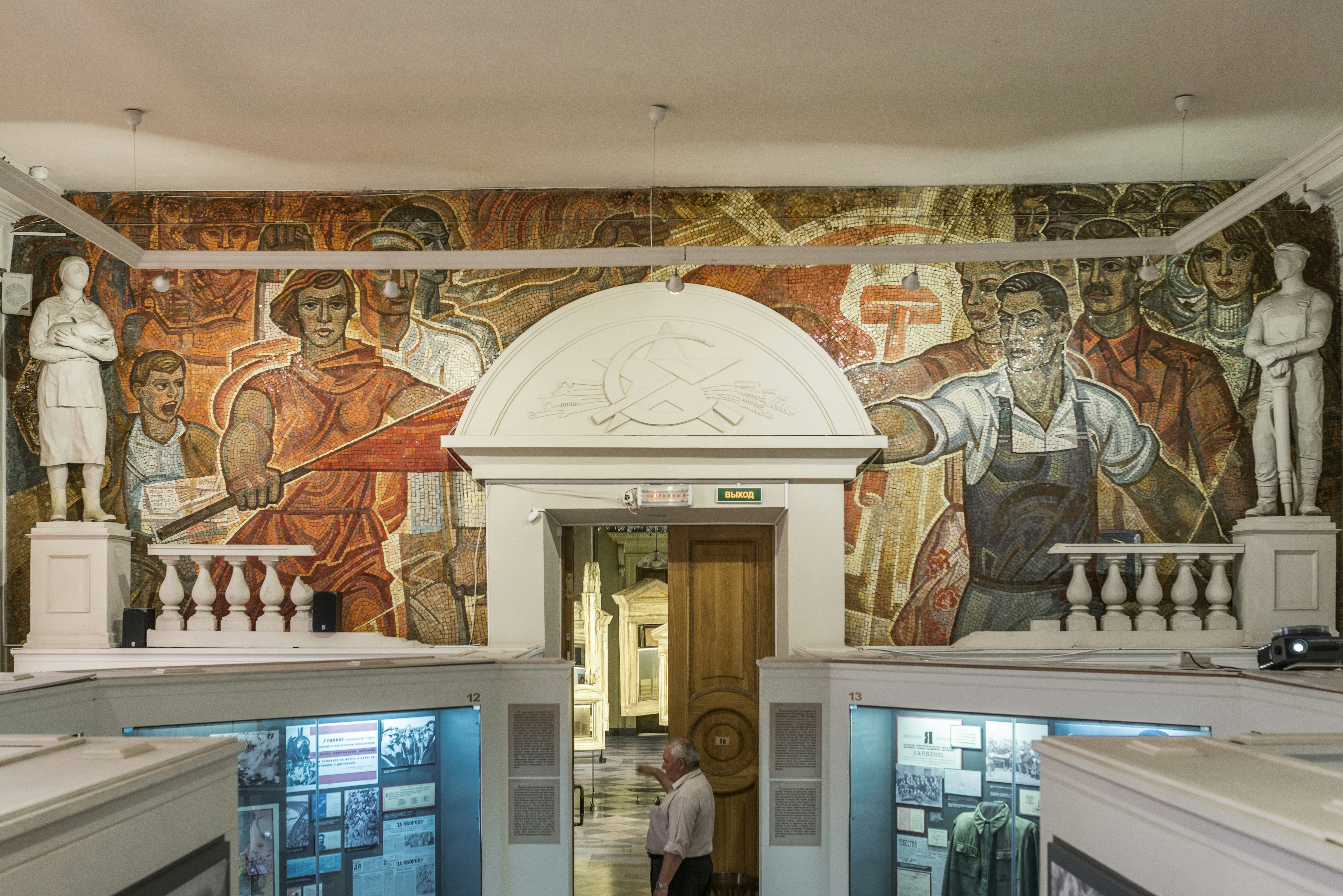 A large mosaic of reds and browns depicting many different people covers a wall. The doorway in the middle of the mosaic has a hammer and sickle emblem above it. Museum displays can be seen below.