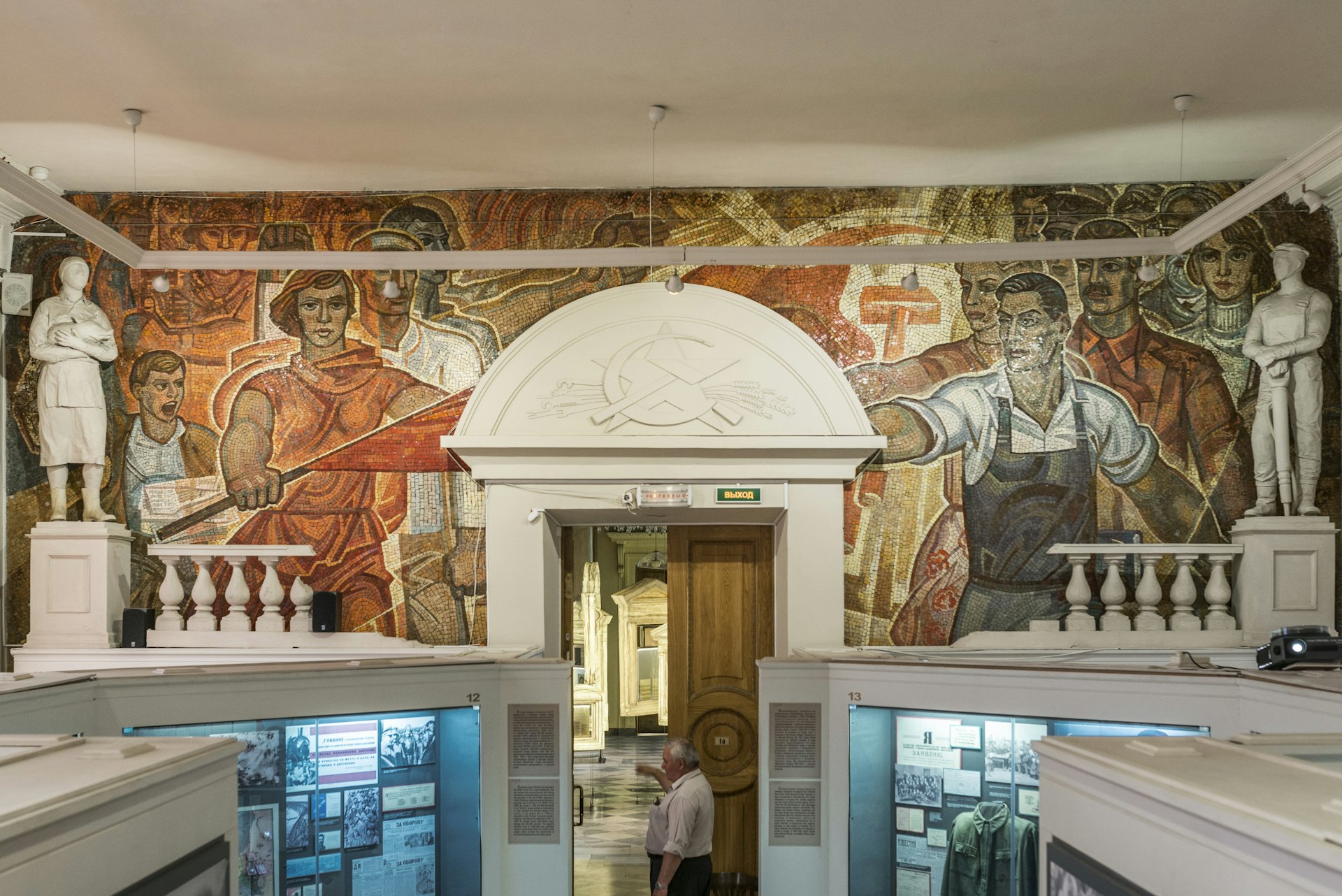 A large mosaic of reds and browns depicting many different people covers a wall. The doorway in the middle of the mosaic has a hammer and sickle emblem above it. Museum displays can be seen below.