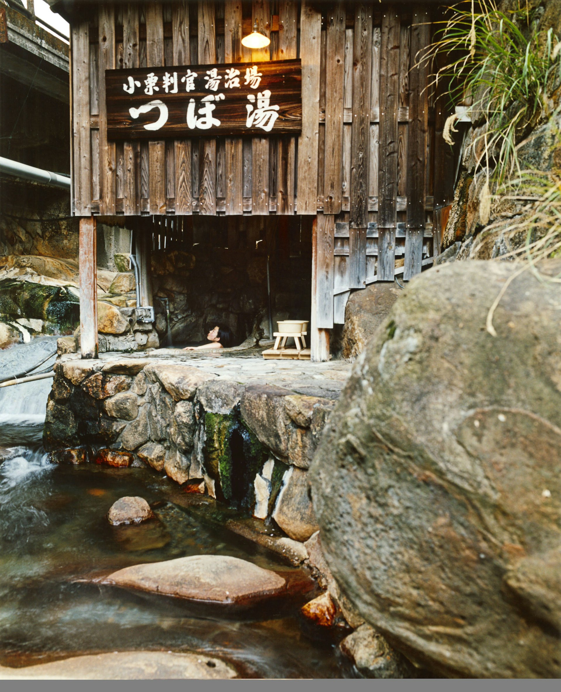 Woman soaking in Tsuboyu Onsen, sheltered by a wooden shack beside river