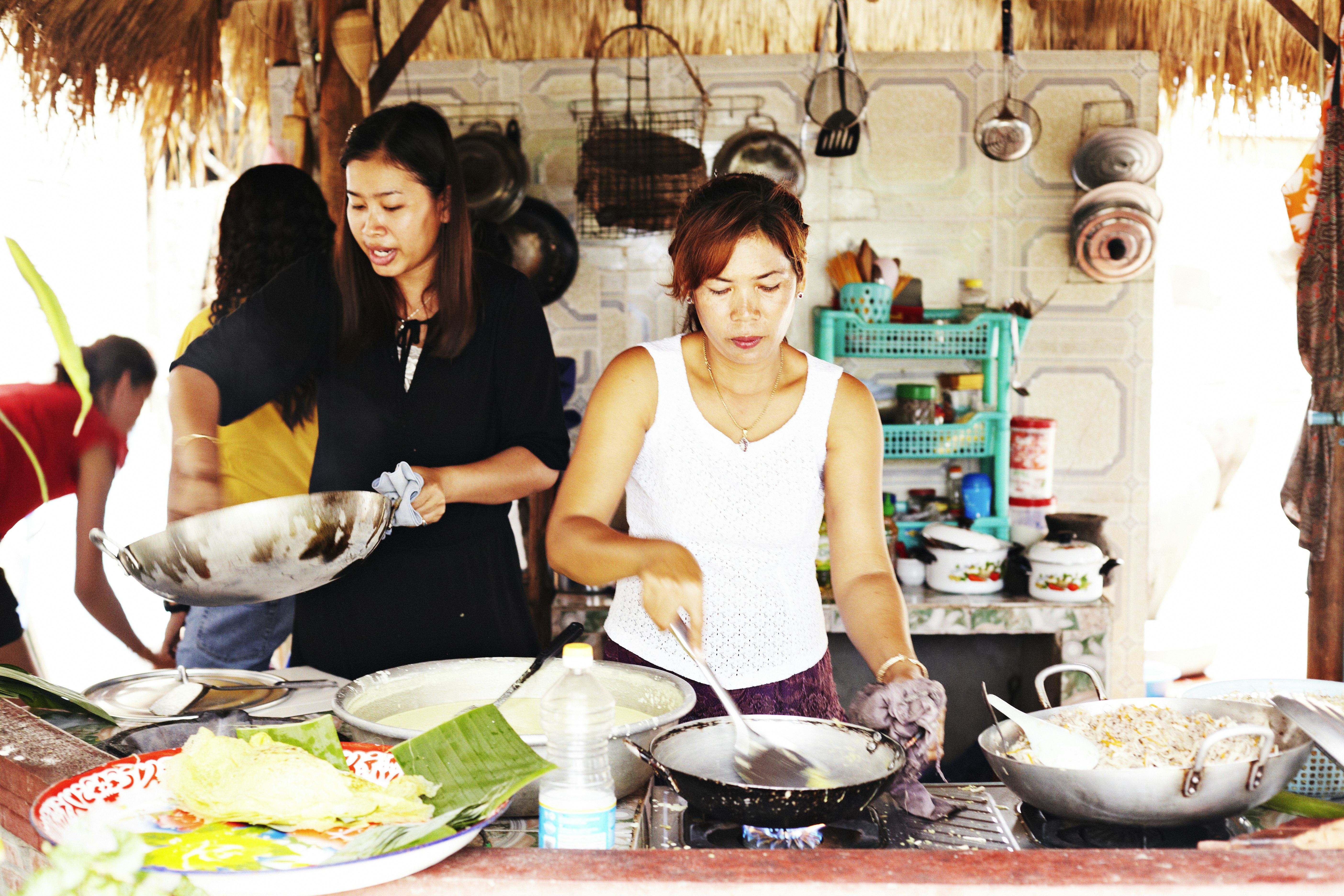 A group of women are cooking up a feast in a homestay. There are many woks on a stove, and behind them there's lots of cookware hanging from the wall, and sitting on a counter.